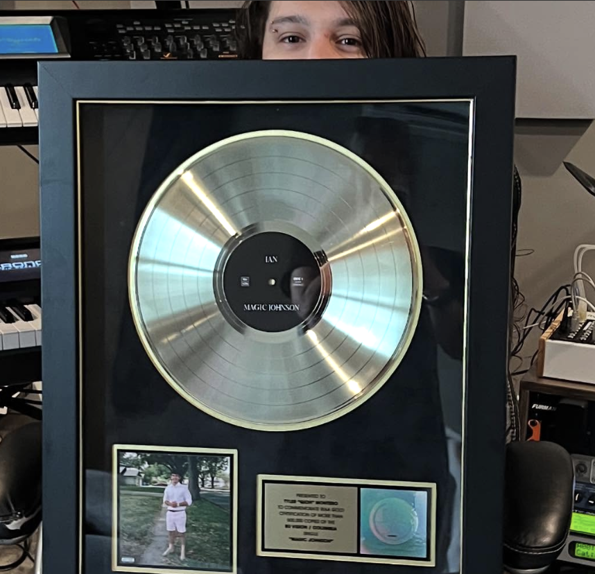 A person peeking over a framed platinum record awarded to Magic Johnson for an album titled 'IAN.' The record is displayed in a black frame with gold and silver accents, and there are two small plaques, one of which features a photo of a man in a park holding a drink, wearing a white shirt and shorts.