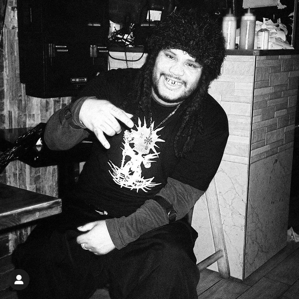 A man with long dreadlocks and a hat, smiling and pointing at his shirt featuring a graphic design, sitting on a wooden chair in an indoor setting with a brick wall and counter in the background.