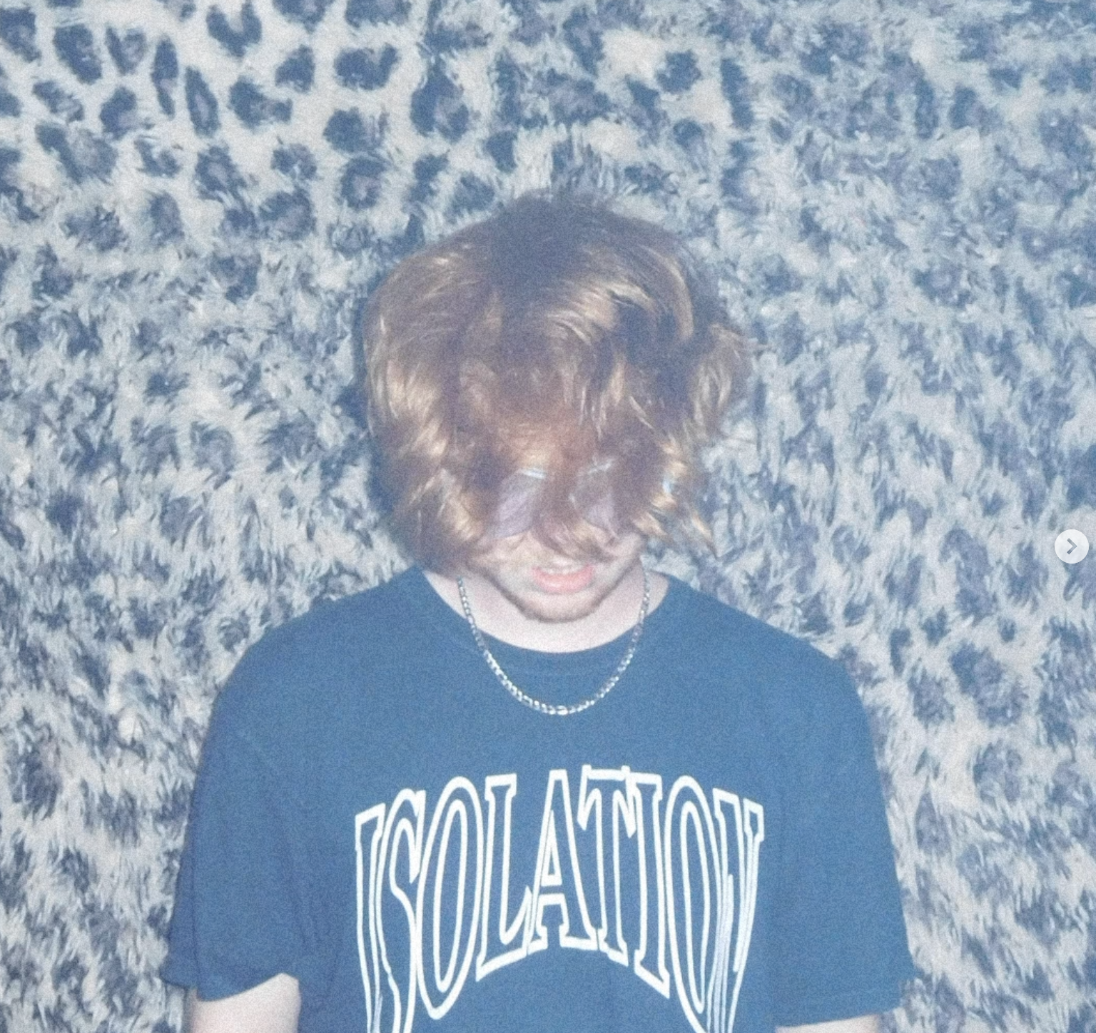 A person with medium-length wavy hair covering their face, wearing black sunglasses, a black T-shirt with white lettering, and a chain necklace, standing in front of a textured, patterned wall.