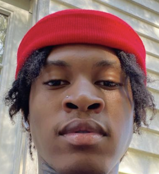 Close-up of a young man wearing a red beanie, earrings, and a nose piercing, outdoors with a metal and wooden siding background.