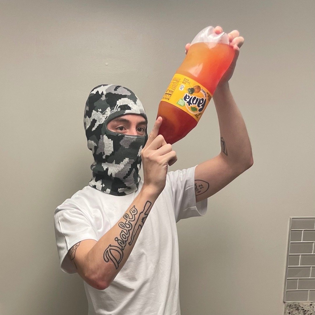 A person wearing a camo face mask and a white t-shirt holding a large plastic bottle of orange Fanta soda.