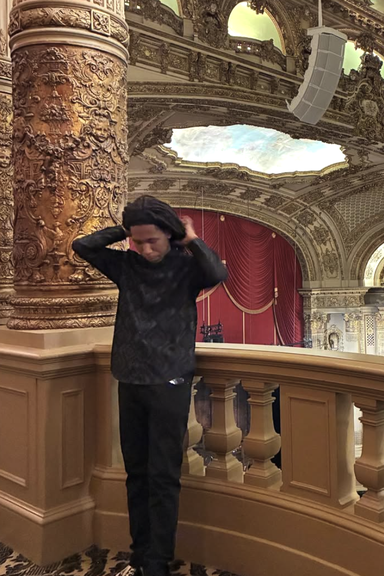 A young man with dark curly hair standing on a balcony in an ornate theater or concert hall, adjusting his hair with both hands. The background features elaborate gold ornamentation, a large carved column, a red curtain, and a painted ceiling with a skylight.