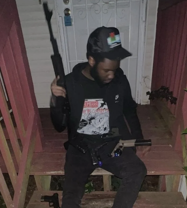 A young man sitting on a wooden porch, holding a rifle in a ready position. He is wearing a black jacket, a black graphic t-shirt, a black baseball cap with a colorful logo, and black pants.