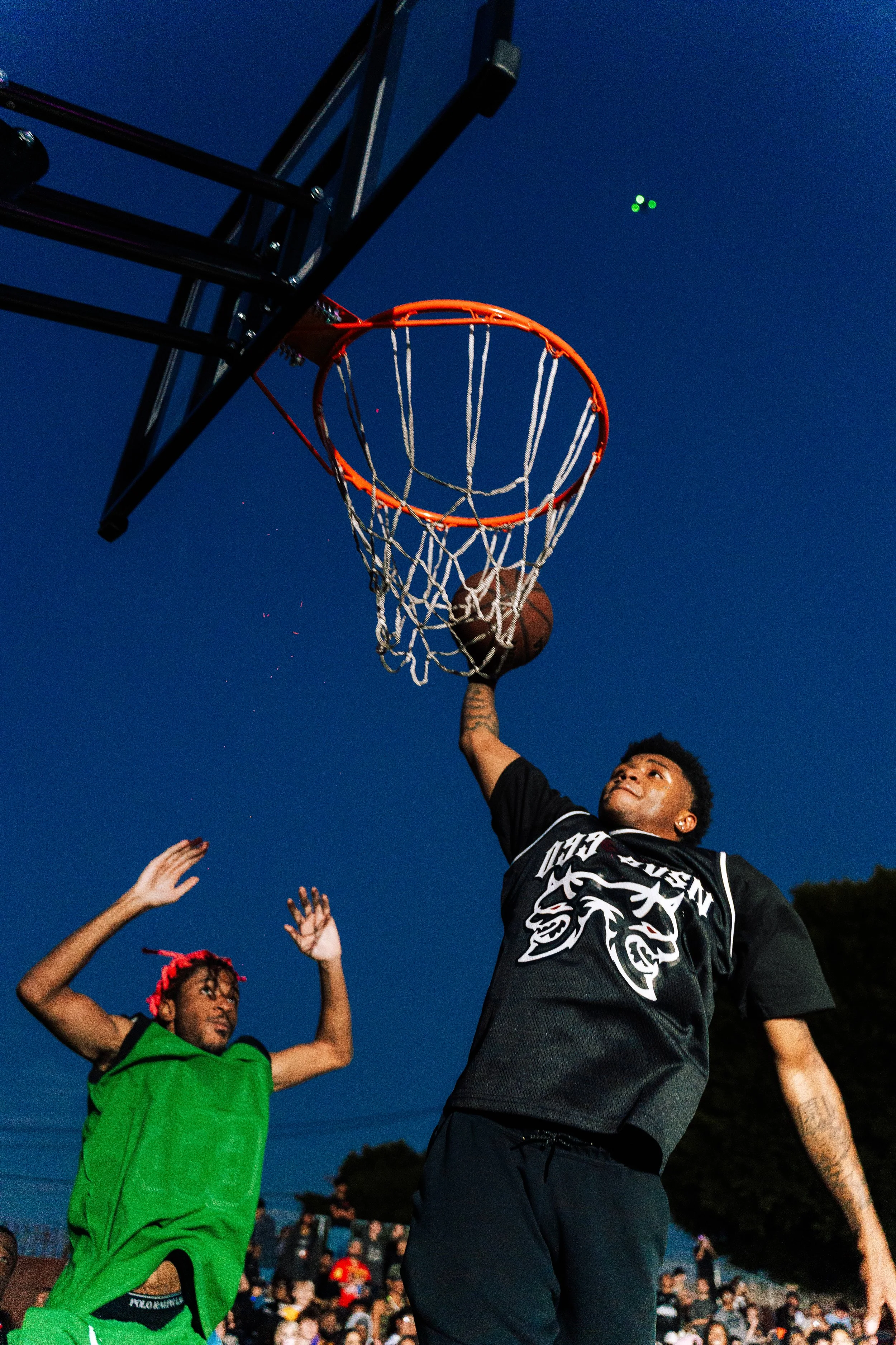A basketball player in black jersey dunking a basketball into the hoop during an outdoor game at dusk, with another player in green attempting to block and spectators in the background.