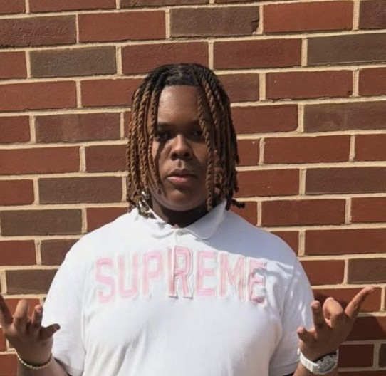 A man wearing a red bucket hat with white patterns, large black sunglasses, a white T-shirt, and earrings, standing against a plain wall.