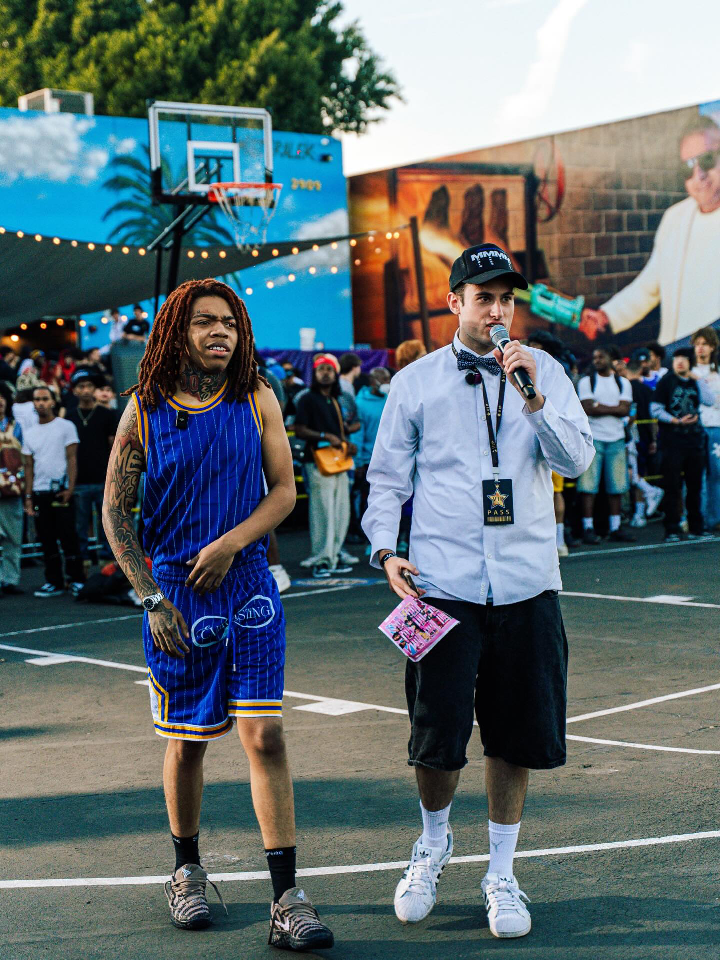 Chuckyy, a rapper, stands on an outdoor basketball court, appearing confused or concerned. Next to them, 42CEO in a white shirt, black shorts, and a cap is speaking into a microphone, holding a pamphlet. A crowd is gathered behind them.