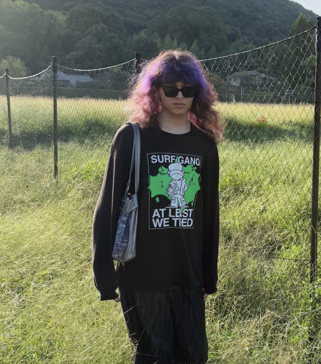 A young woman with curly purple and pink hair wearing sunglasses, a black graphic shirt with text 'SURF GANG AT LEAST WE TIED', and black pants, standing outdoors near a chain-link fence on a sunny day with a grassy field and hills in the background.