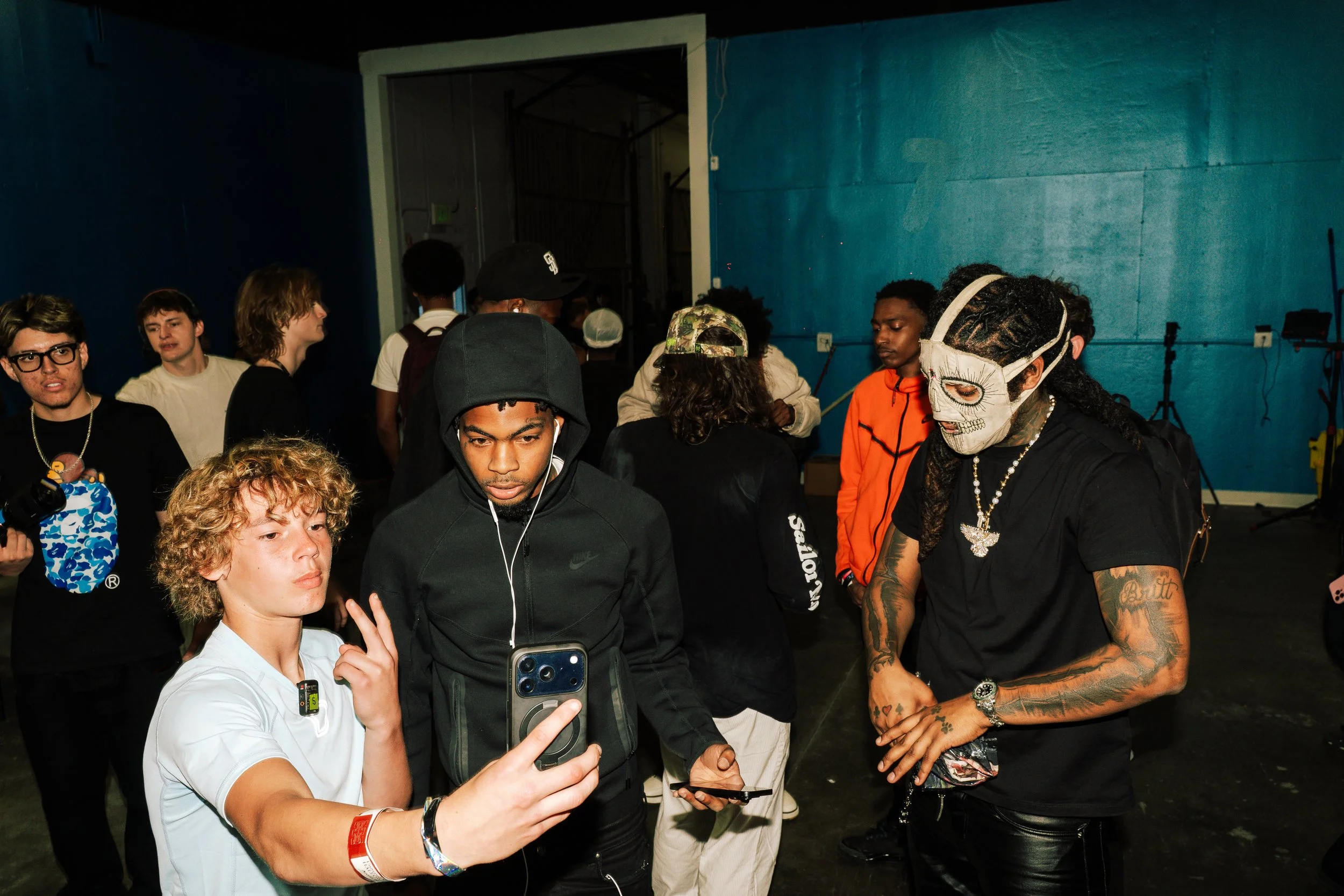 A group of young people at an indoor event, some taking photos or looking at their phones, with a blue wall in the background.