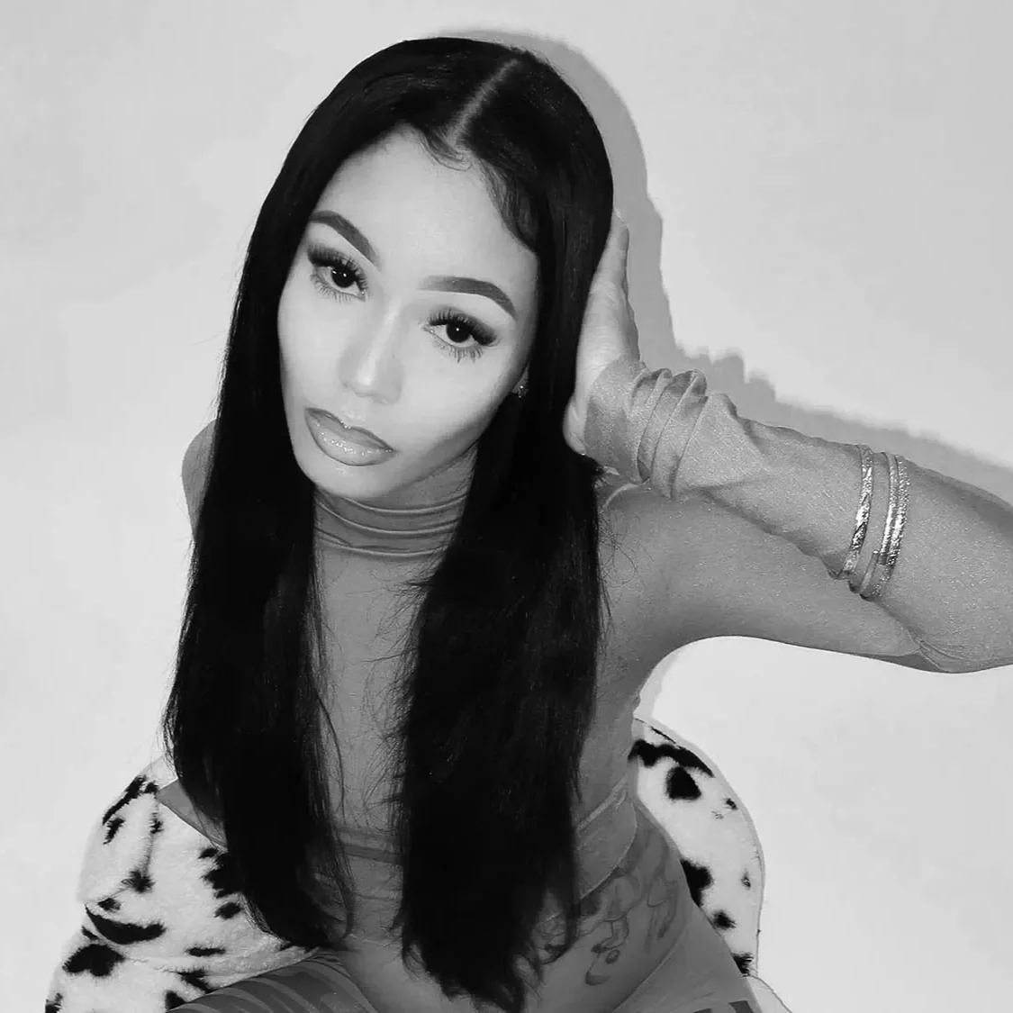 Black and white photo of a woman with long, dark hair, wearing makeup, a turtleneck, and sitting in a leopard print outfit, with one hand touching her head and wearing bracelets.