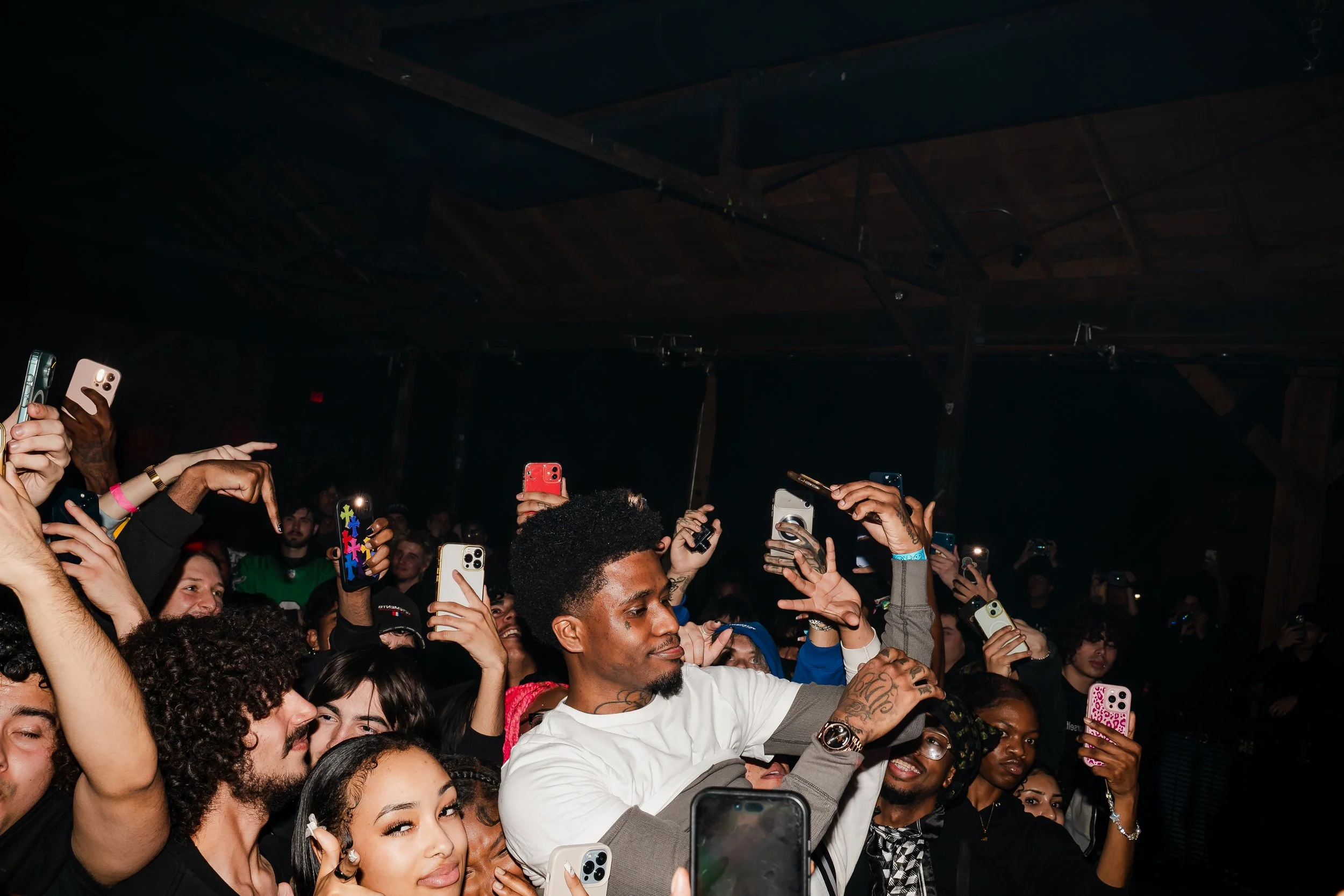 A crowd of people at a concert or party, many holding up smartphones, with a man in a white shirt in the center taking a picture or video.