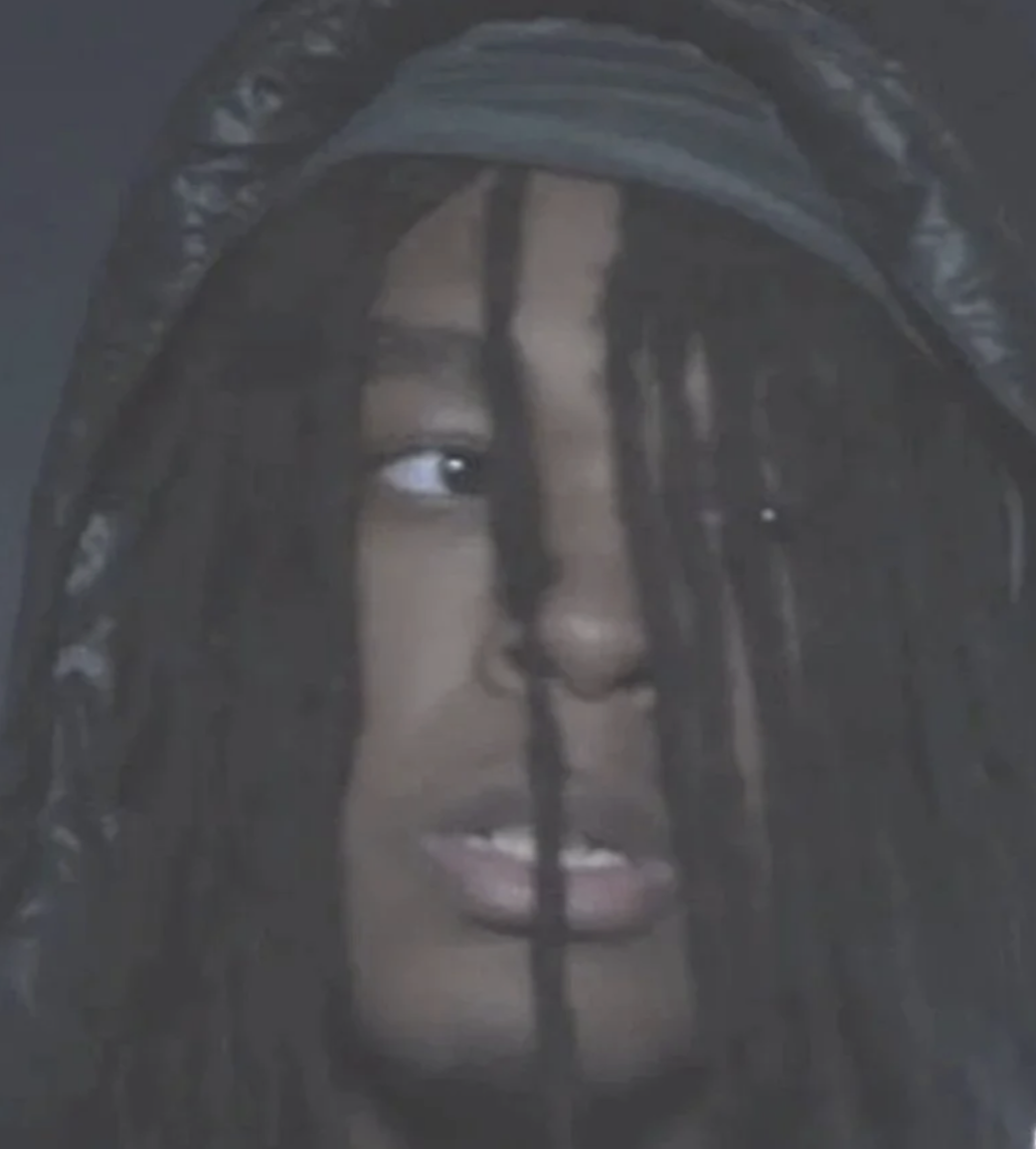 Close-up of a person's face with dreadlocks, wearing a hood, and looking to the side.