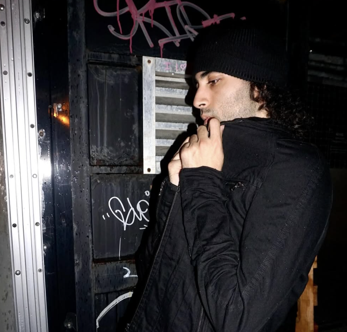 A young man in a black beanie and black jacket standing near a graffitied wall, with his hand near his face, looking thoughtful.