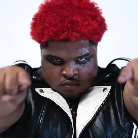 Person with bright red curly hair looking intently at the camera, wearing a black leather jacket with the zipper partially open, and pointing fists towards the camera.