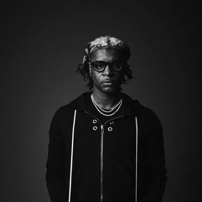 A black-and-white portrait of a person with glasses and short, curly hair, wearing a hoodie and layered necklaces against a dark background.