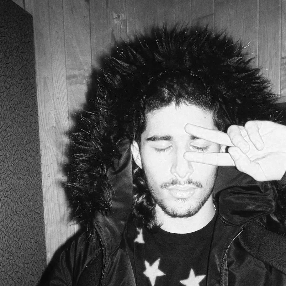 A young man with long hair, a mustache, and a beard, wearing a black jacket with a fur-lined hood, making a peace sign with his fingers near his face and closing his eyes.