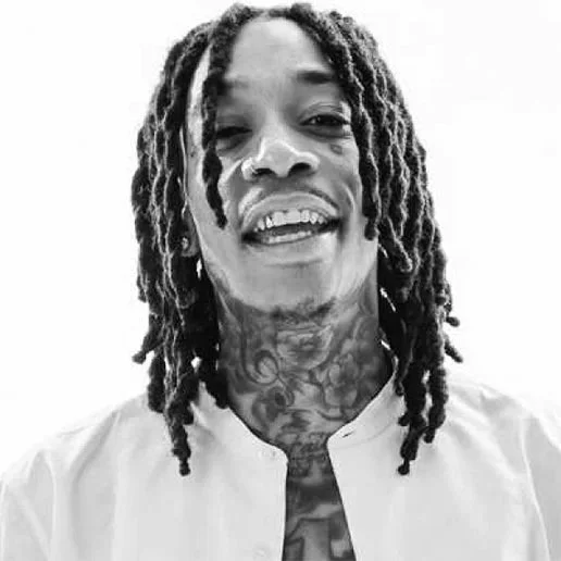 Black and white photo of a smiling man with dreadlocks, tattoos on neck and chest, wearing a light-colored shirt, against a plain background.