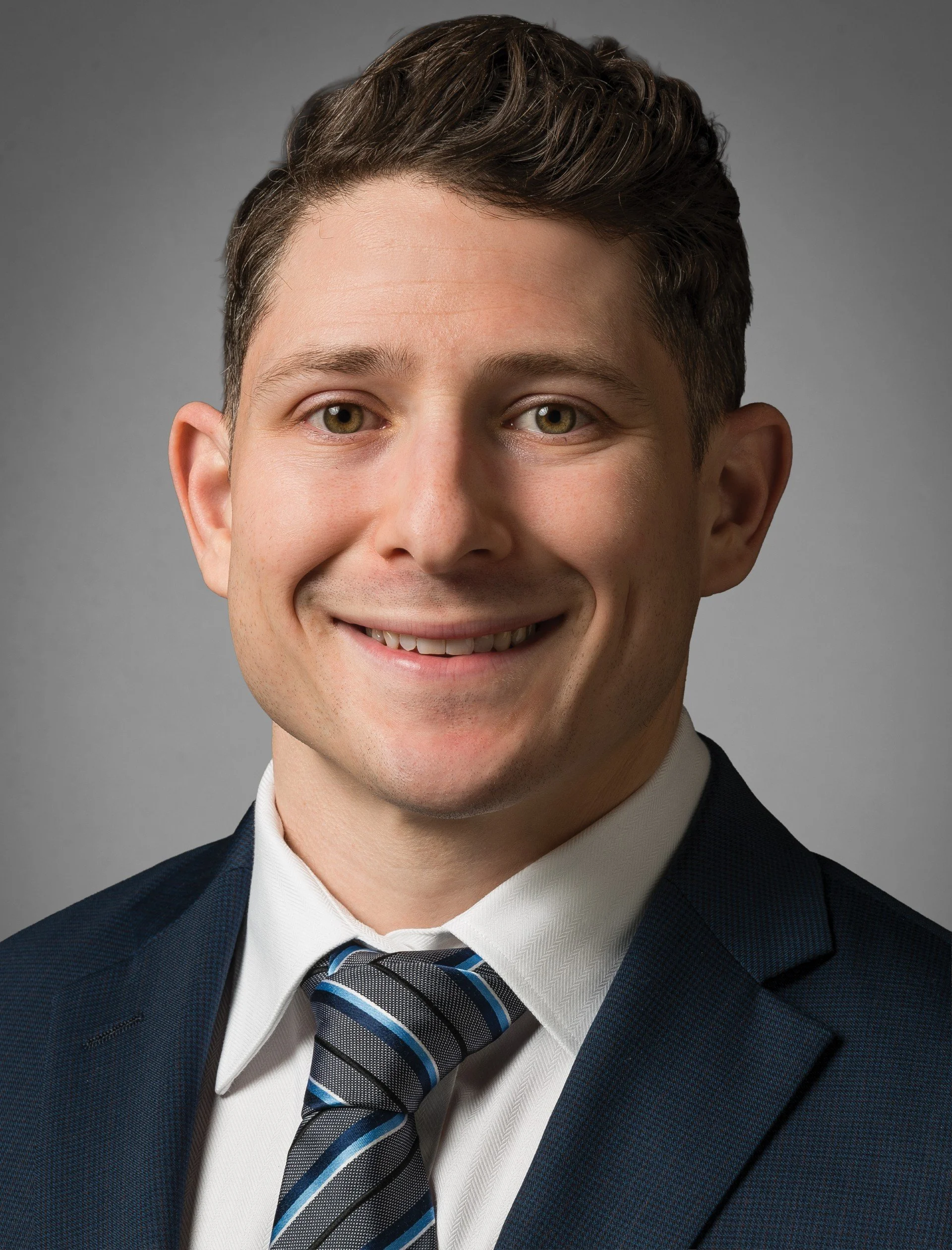 Close-up of a young man in formal business attire, smiling, with brown hair and green eyes, wearing a dark blue suit, white shirt, and a striped blue and black tie against a gray background.