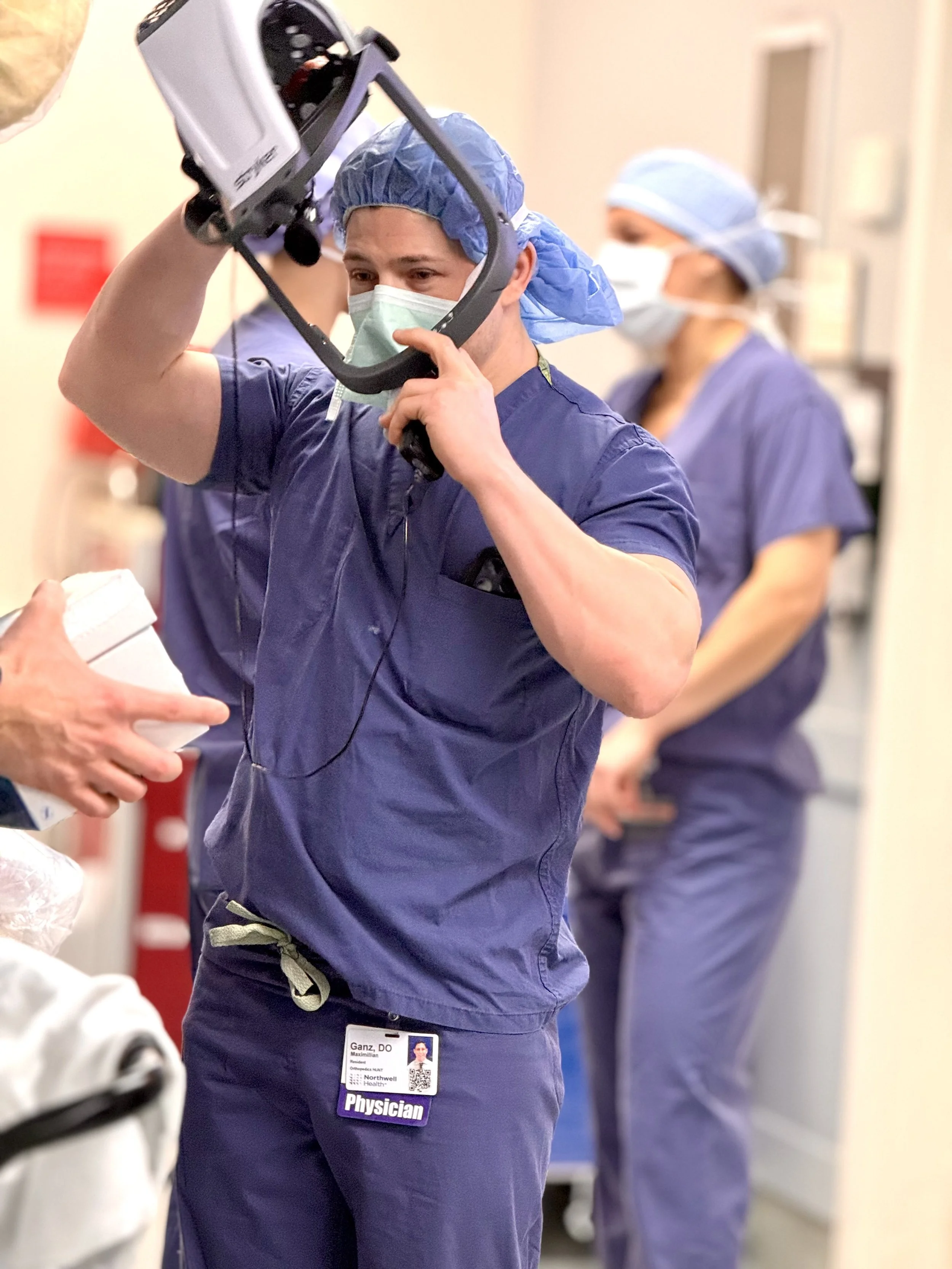 A total joint arthroplasty surgeon, Dr. Maximillian P Ganz, healthcare worker in blue scrubs and a surgical mask holding an X-ray imaging device, with other medical staff in the background.