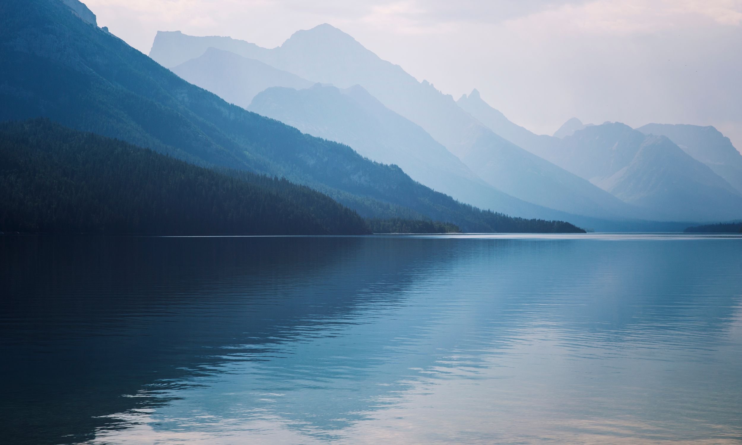 A serene mountain lake with calm waters reflecting the surrounding forested mountains under an overcast sky.