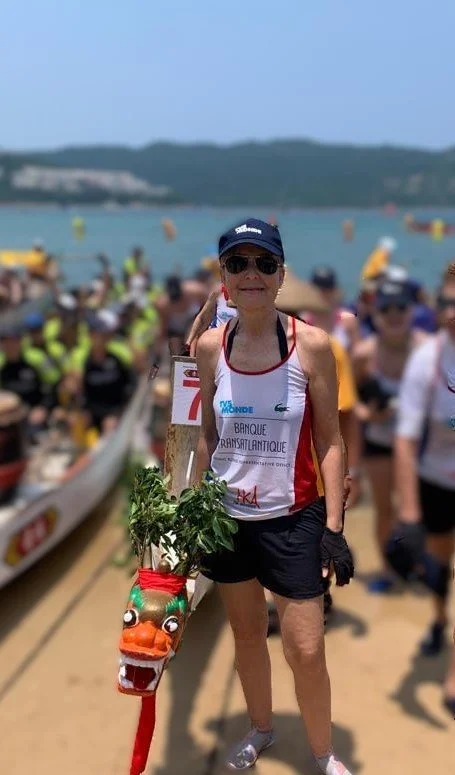 An elderly woman standing on a beach in front of a crowd during a boat event. She is wearing a tank top, shorts, sunglasses, and a cap, and is holding a decorative dragon boat figure with greenery and a dragon face.