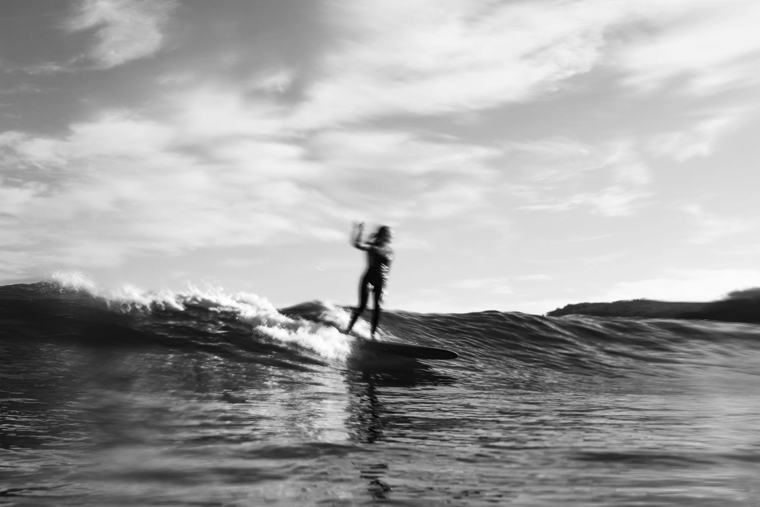 A person surfing on a wave in the ocean with a cloudy sky in the background, shown in black and white.