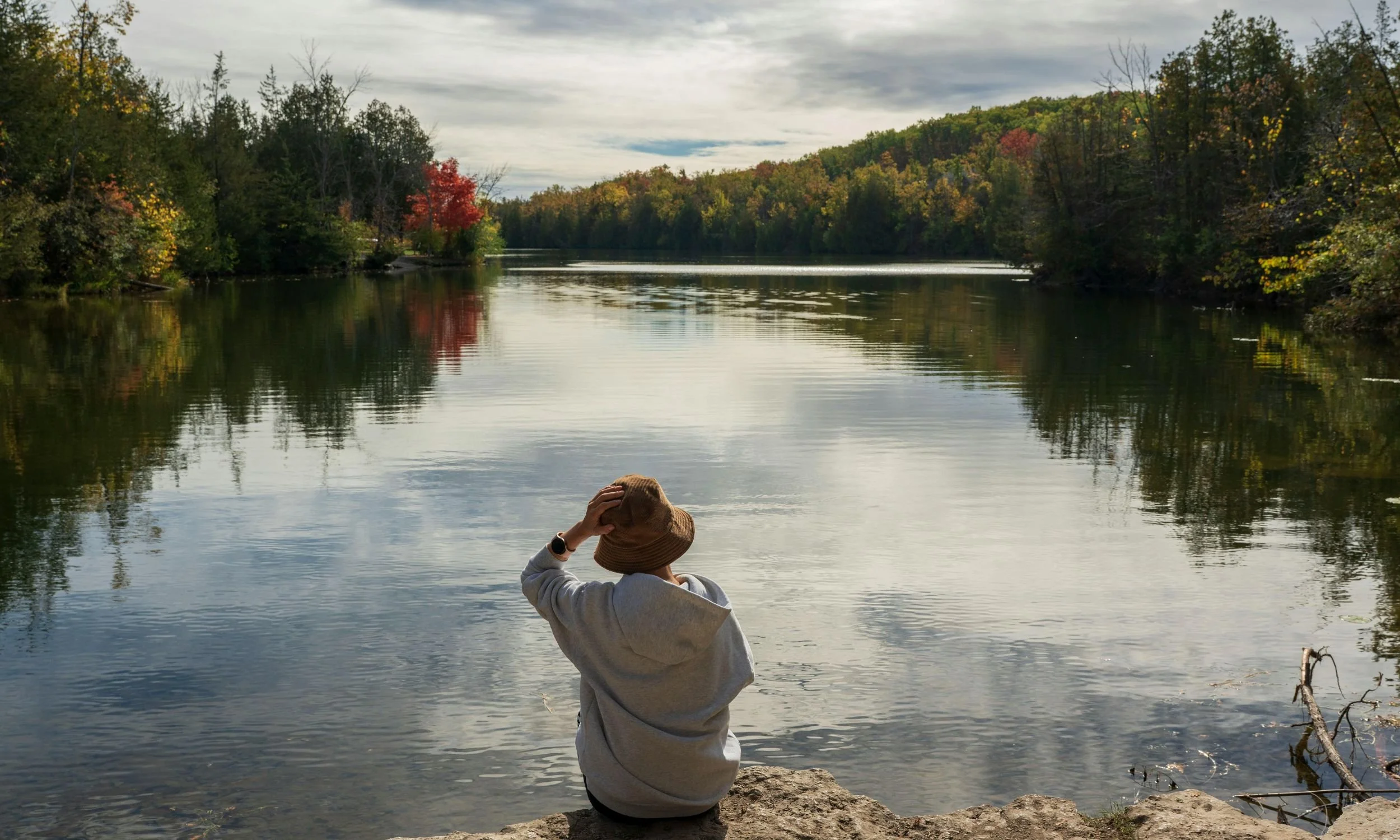 Person sitting on rocks by a calm river or lake, wearing a hat and hoodie, surrounded by autumn trees with colorful leaves, under a partly cloudy sky.