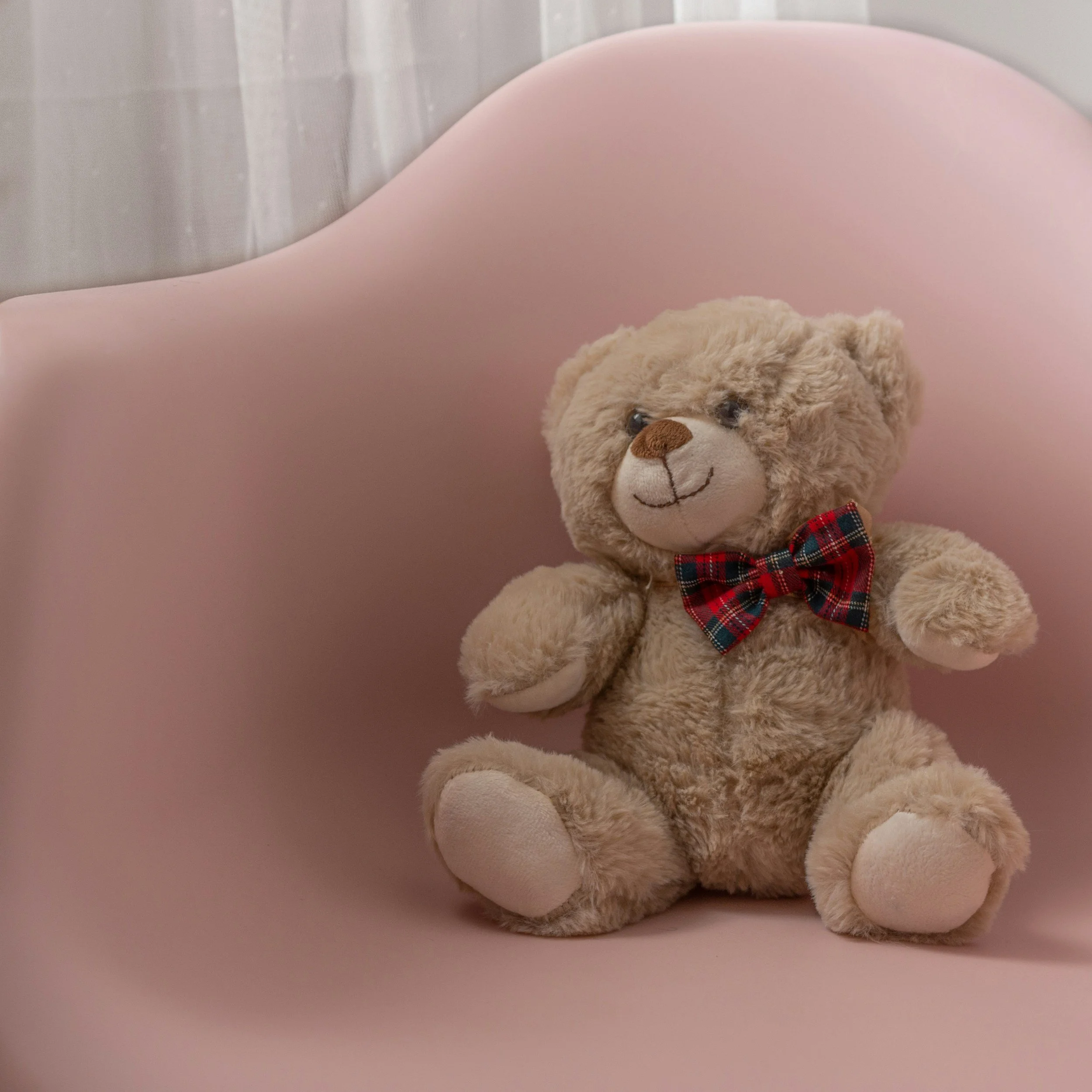 A plush teddy bear wearing a red plaid bowtie sitting on a pink chair with cream-colored curtains in the background.