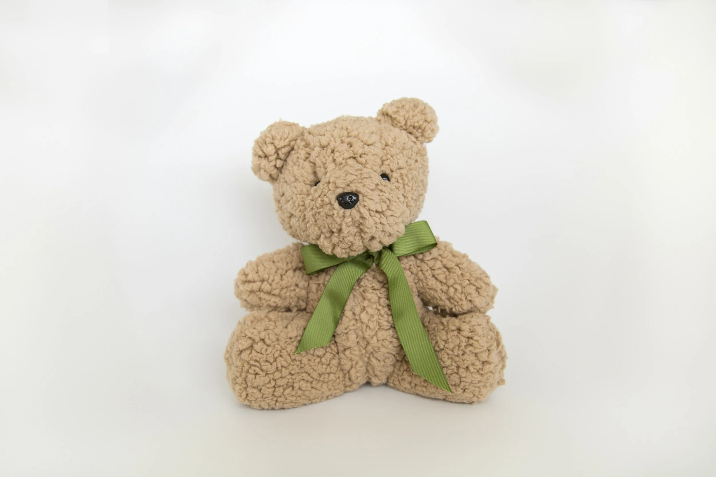 A plush teddy bear with beige, curly fur, black eyes, a black nose, and a green ribbon tied around its neck, sitting on a plain white background.