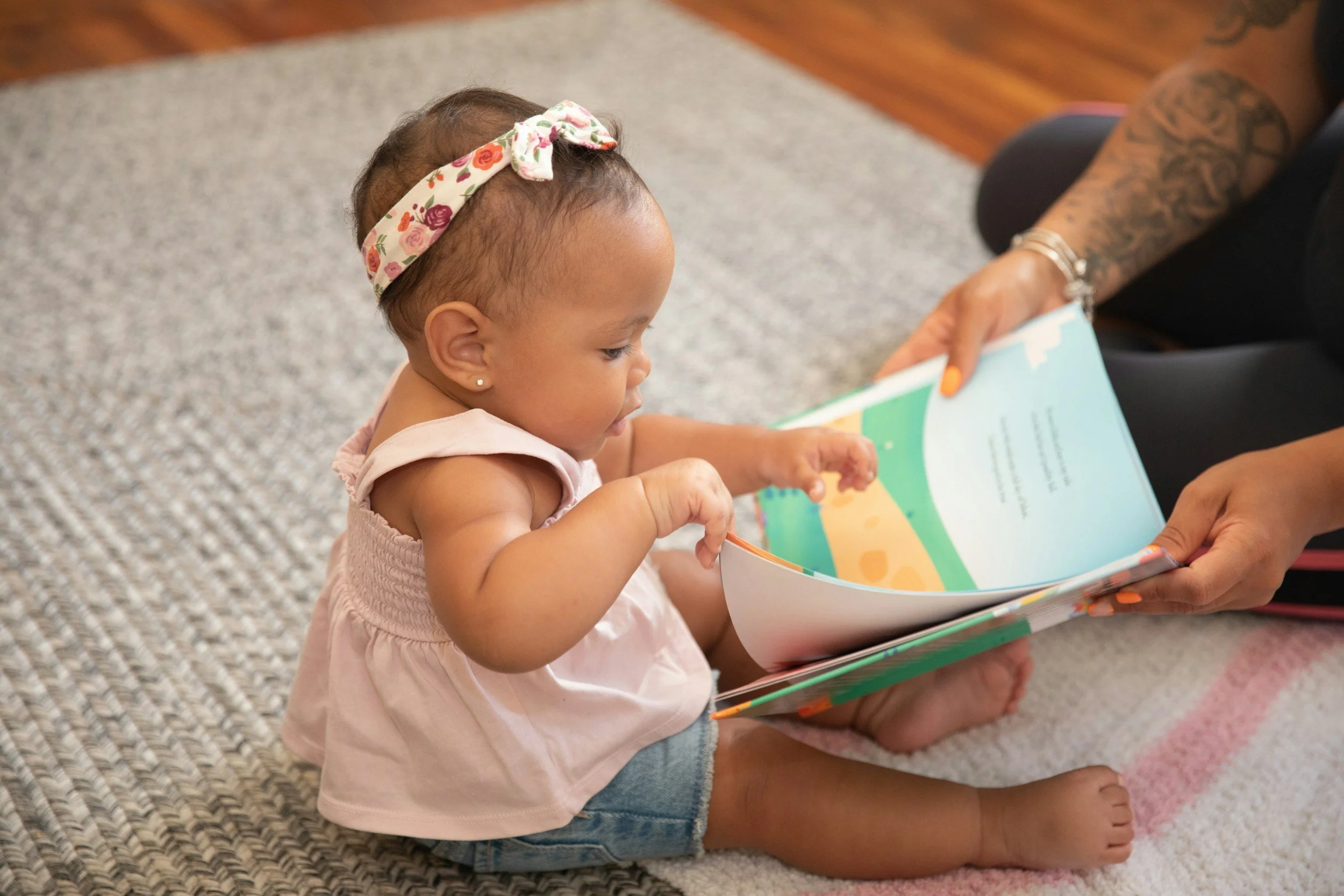 Young girl sitting on a carpet, being read a colorful children's book by an adult with tattoos on their arm.