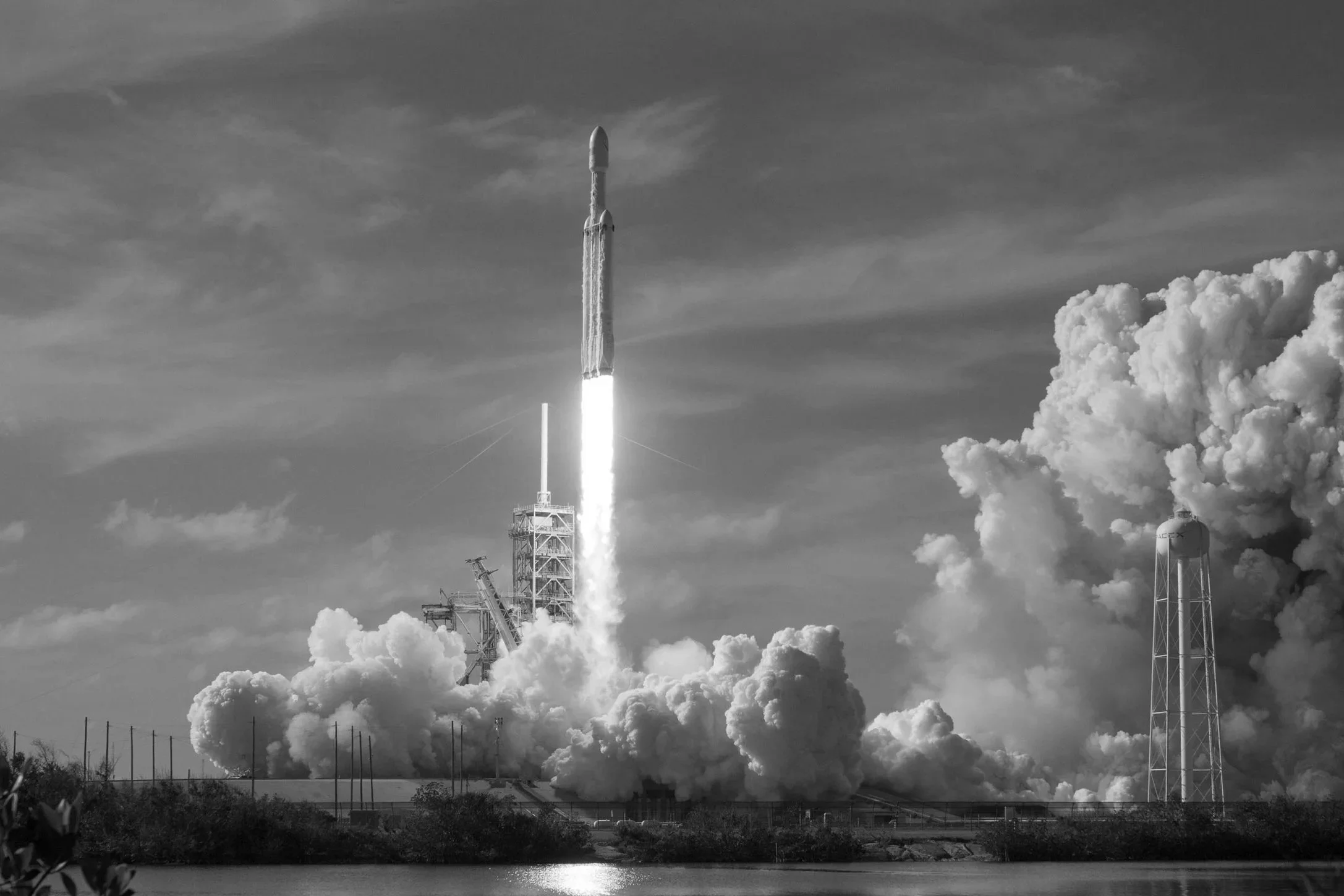 Black and white photo of a rocket launching with a fiery exhaust plume, surrounded by clouds of smoke and steam on the ground, near a water body.