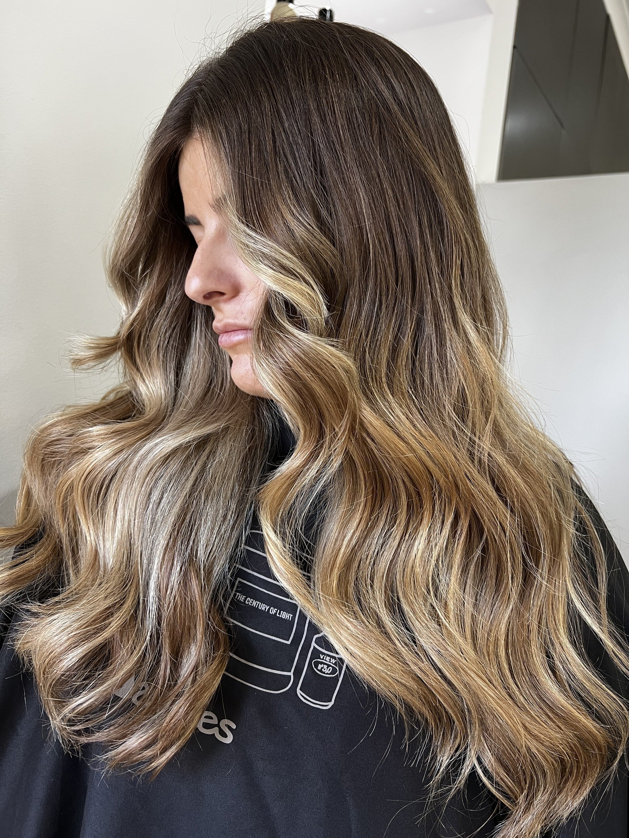 Woman with long, wavy hair featuring a balayage color transition from dark brown at the roots to blonde at the ends, sitting in a salon chair.