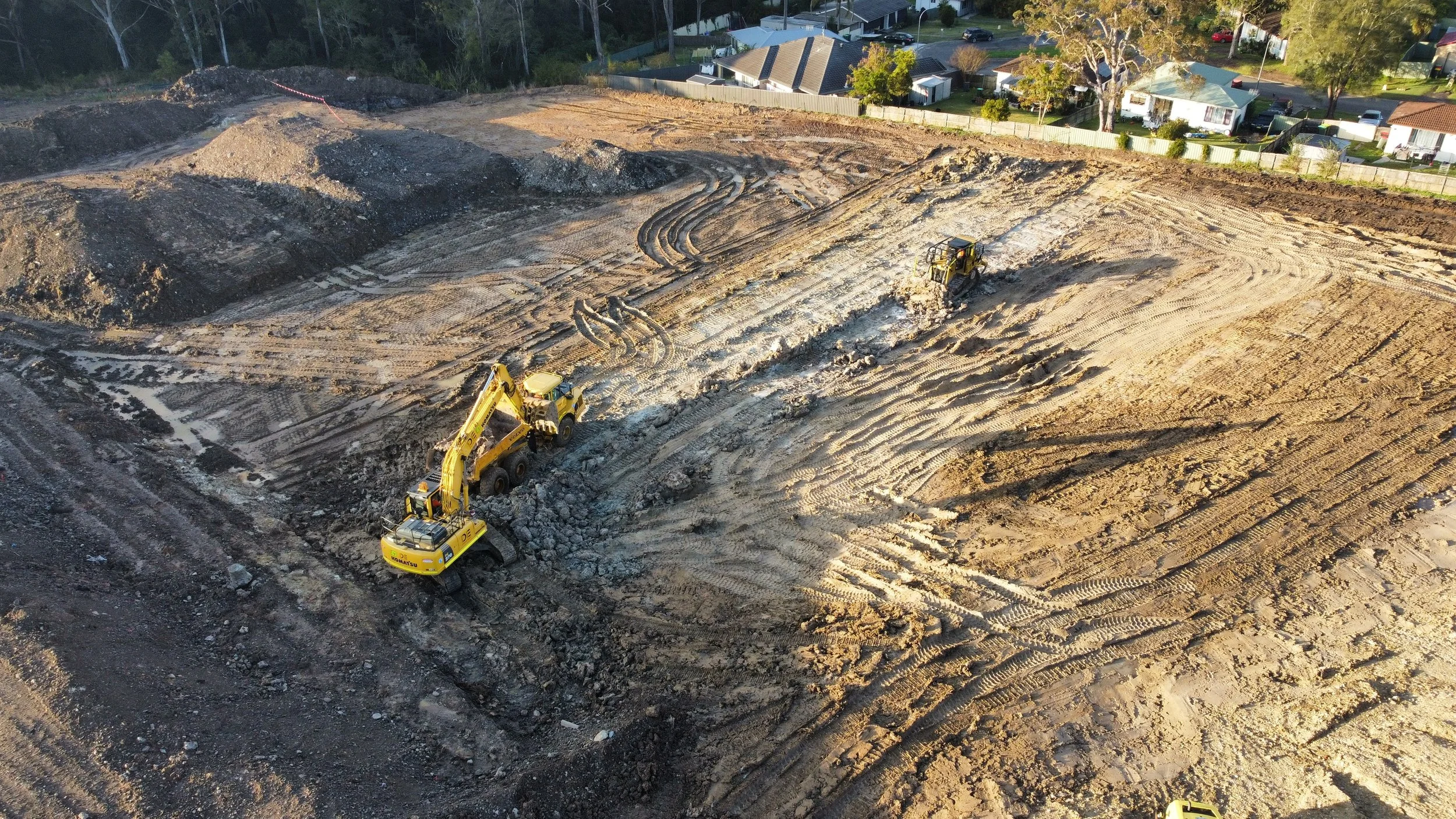 Construction site with two yellow excavators working on leveling the ground, surrounded by dirt and tire tracks, with residential houses and trees in the background.