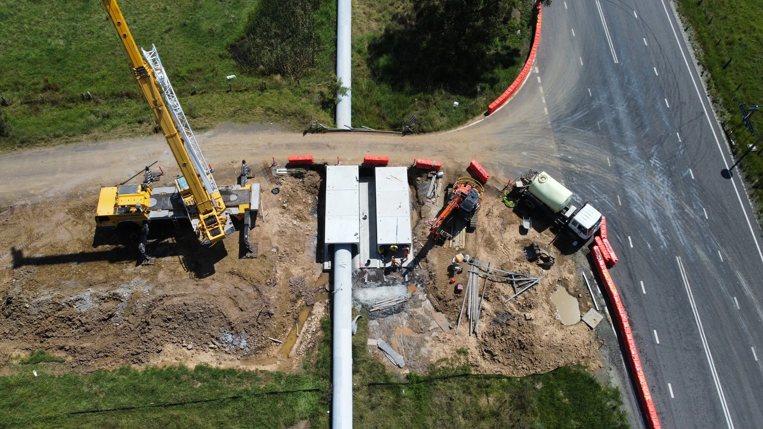 Construction site where workers are installing a large pipe under a road with heavy machinery and safety barriers.
