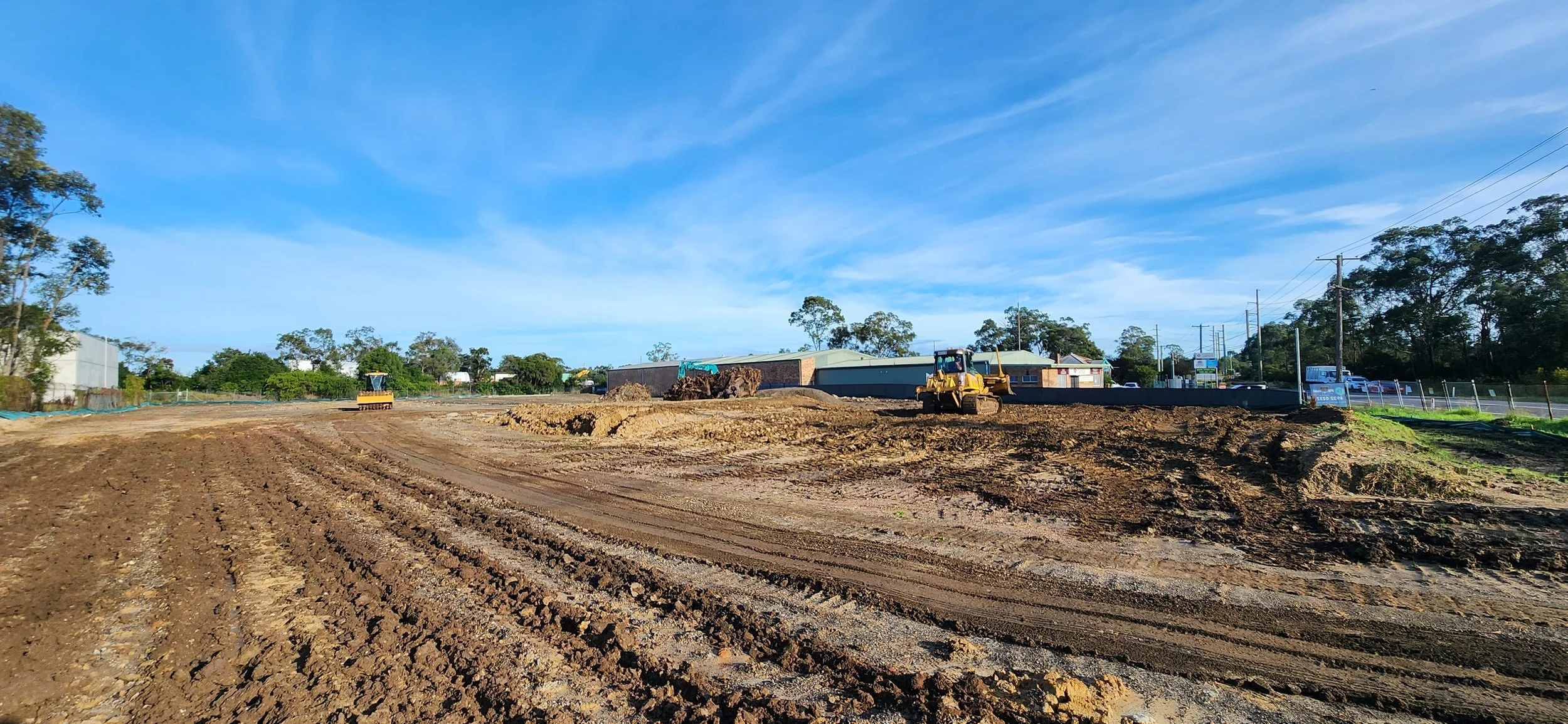 Construction site with heavy machinery on muddy ground, cleared and leveled land, with buildings and power lines in the background under a blue sky with scattered clouds.