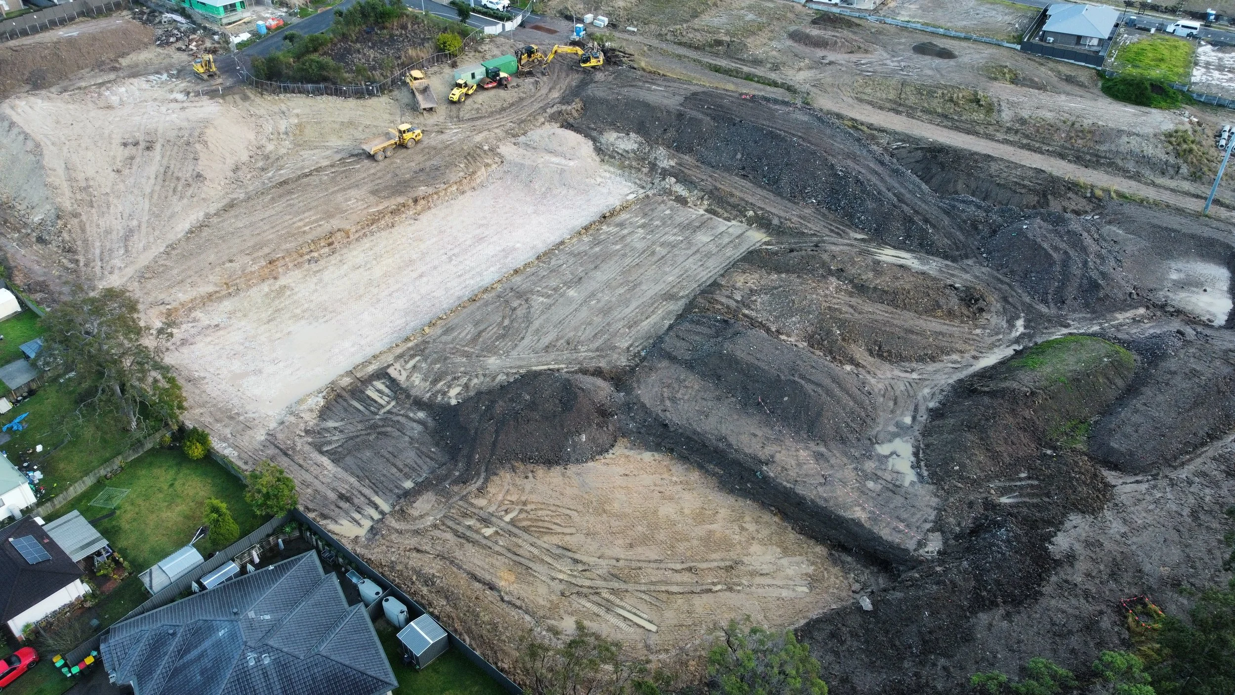 Aerial view of a construction site with cleared land, dirt mounds, and construction machinery, surrounded by residential houses and green yards.