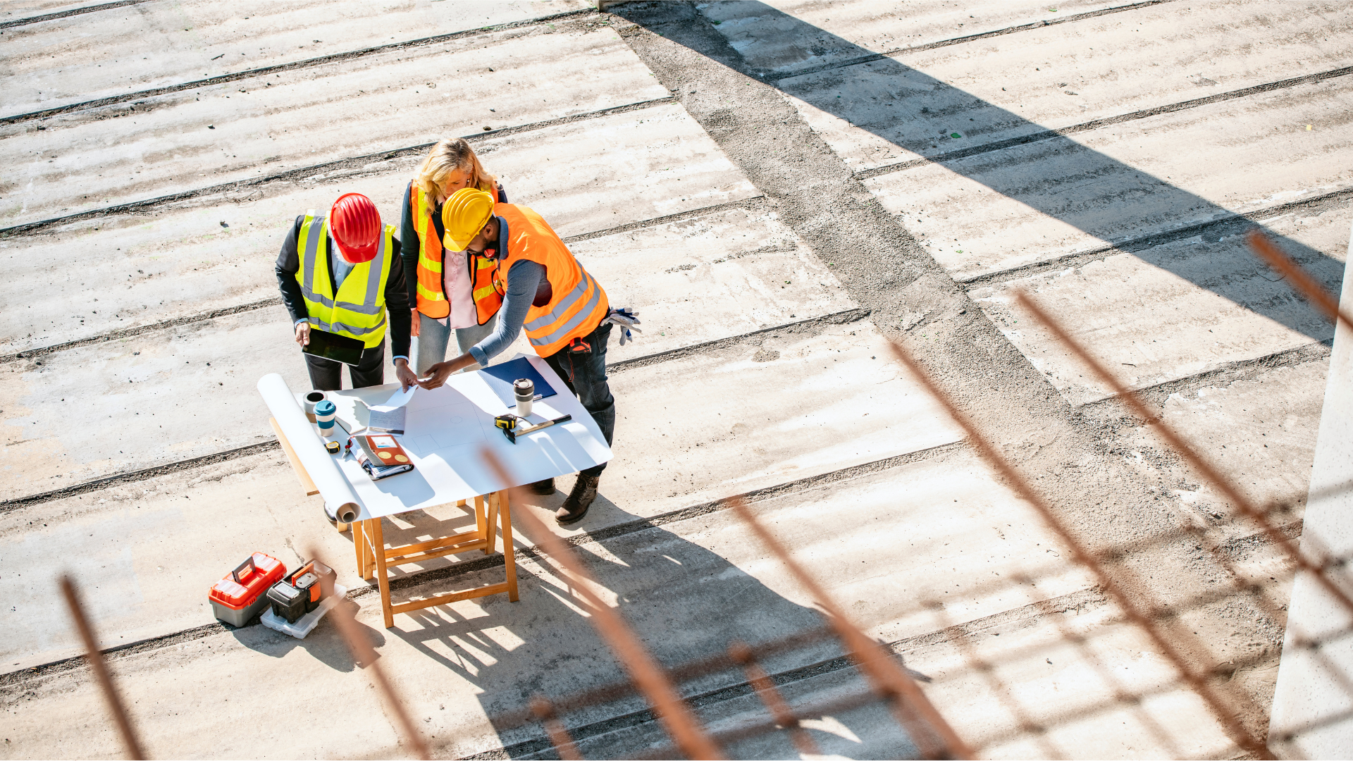Construction workers wearing hard hats and reflective vests discussing plans on a construction site with a table and tools.