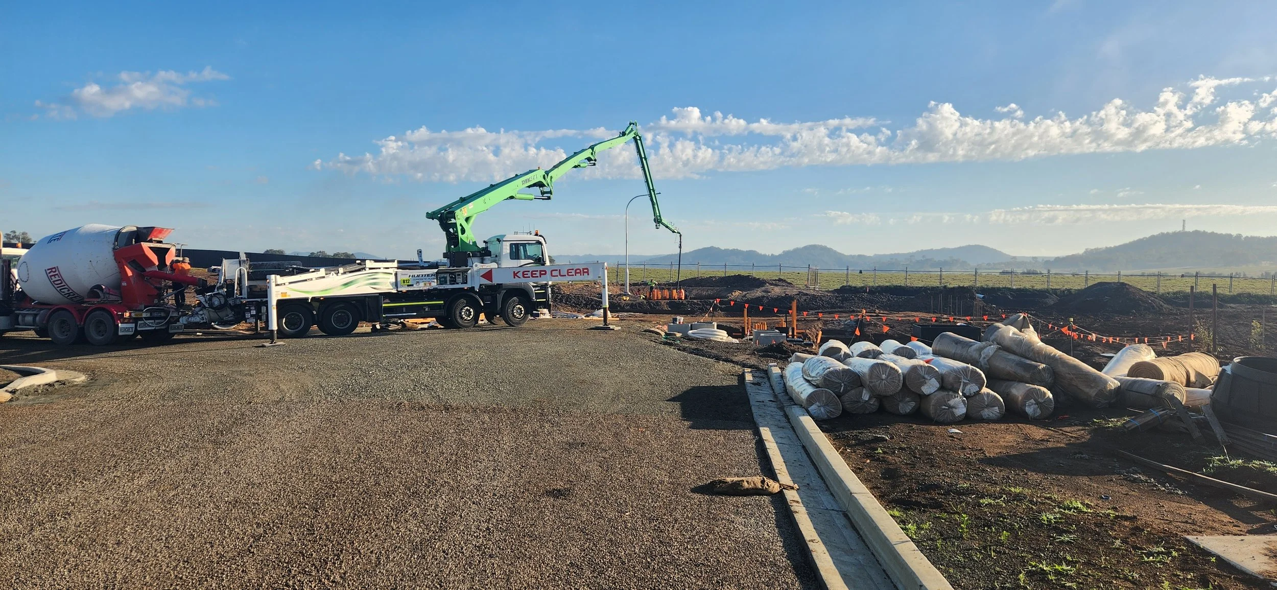 Construction site with a concrete mixer truck, a crane truck, and piles of rolled-up materials, set against a scenic landscape with rolling hills and a blue sky.
