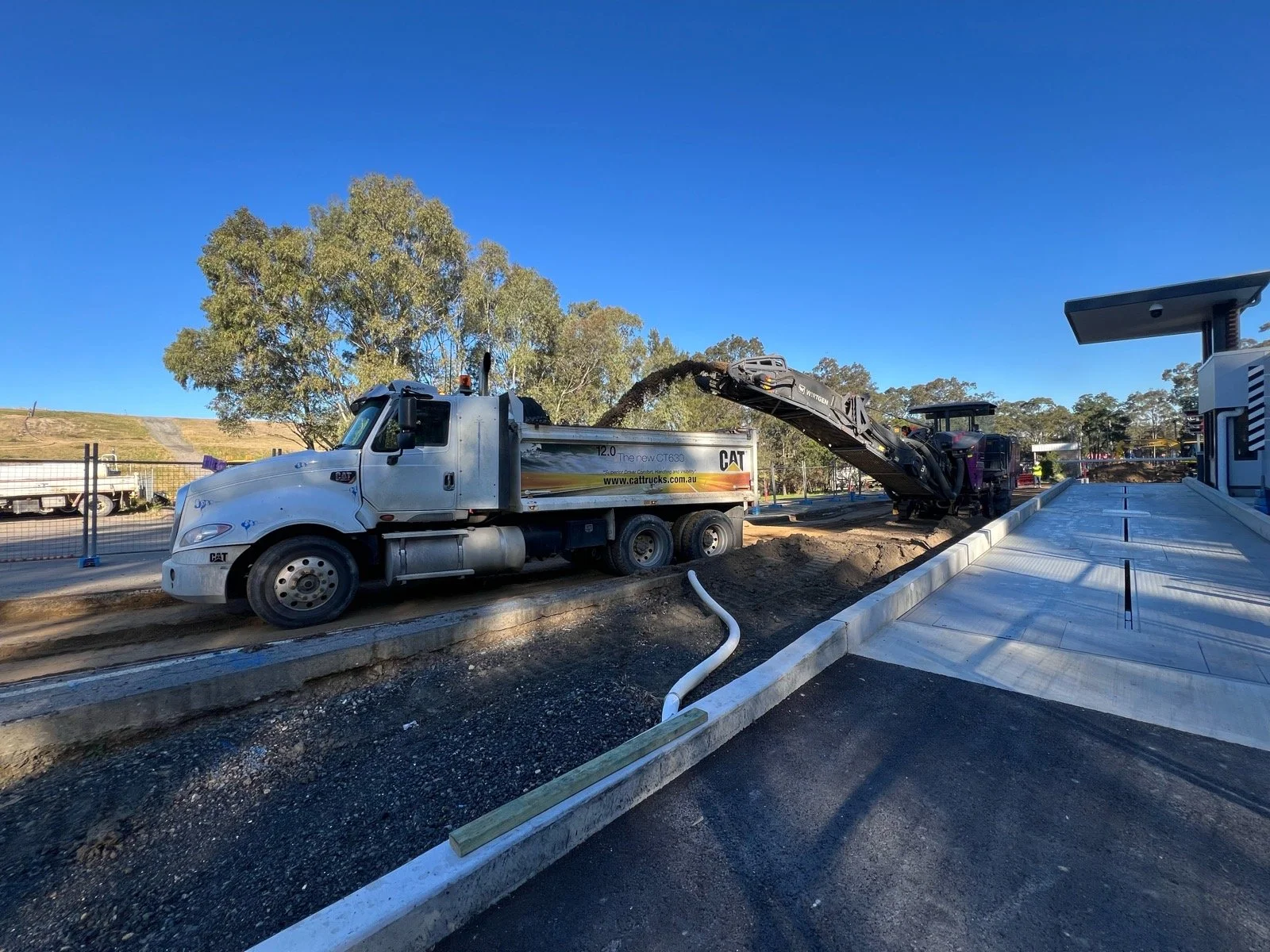 Construction site with a white Caterpillar truck and an excavator transferring dirt, near a sidewalk and trees in the background under a clear blue sky.