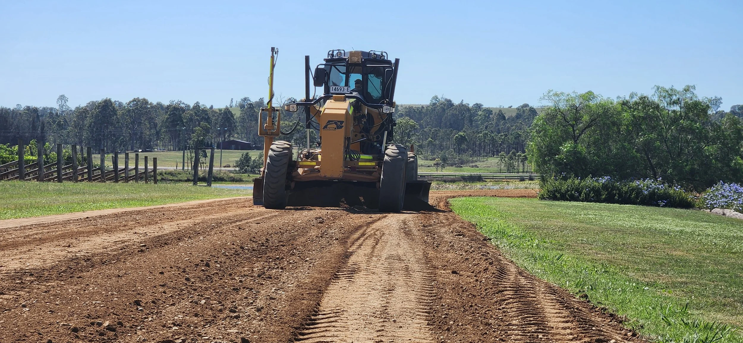 A bulldozer working on a dirt path in a rural landscape with trees and flowers in the background.