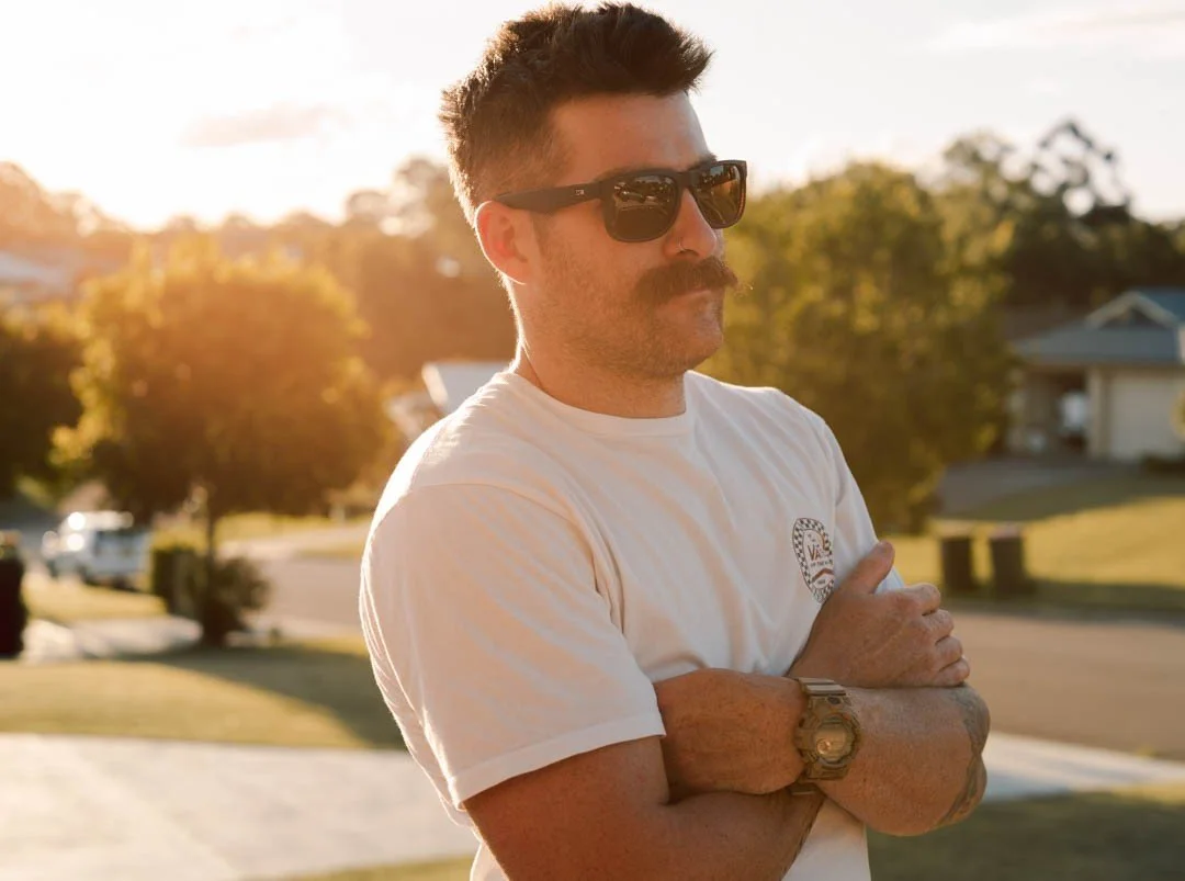 A man with dark hair, sunglasses, and a mustache wearing a white t-shirt with rolled-up sleeves, standing outdoors during golden hour with trees and houses in the background.