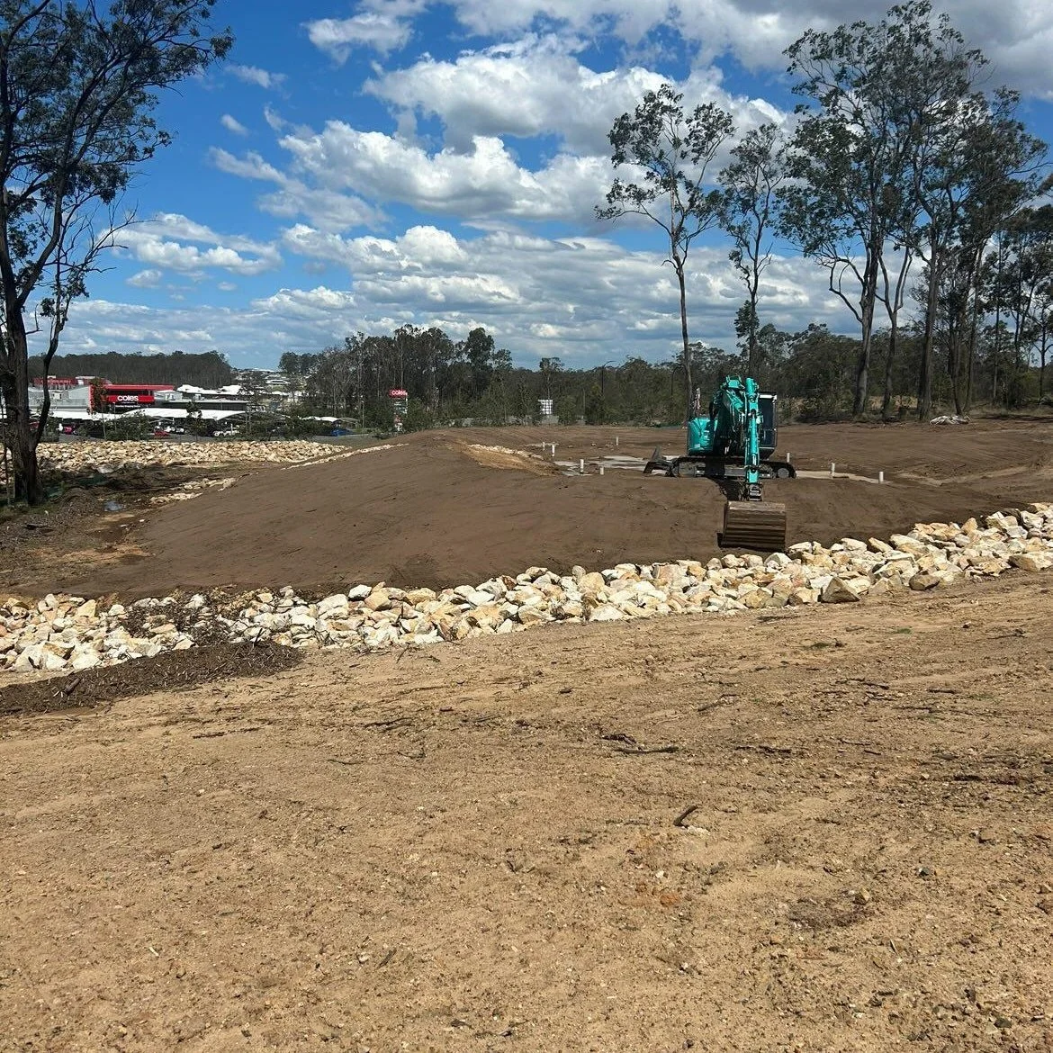 Construction site with a small blue excavator on dirt, surrounded by rocks and trees under a partly cloudy sky.