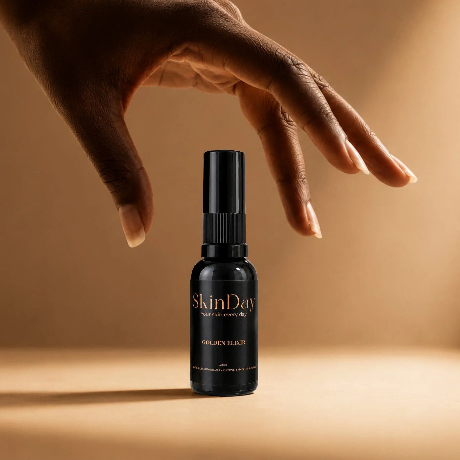 A black spray bottle labeled 'Skin Day,' with the tagline 'Your skin every day,' and the text 'GOLDEN ELIXIR' at the bottom. A hand with dark skin and well-manicured nails is reaching toward the spray nozzle against a neutral background.