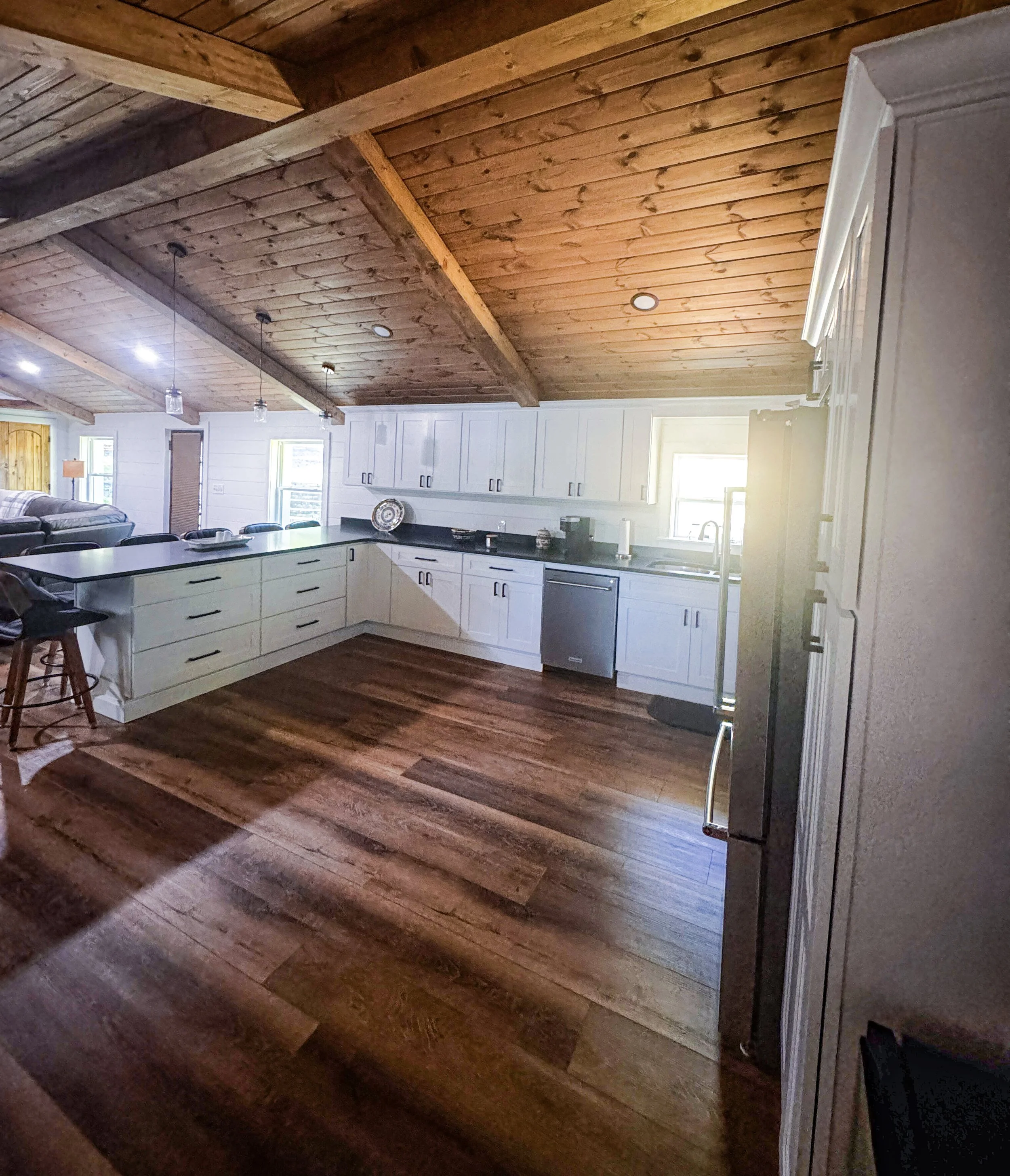 A kitchen with white cabinets, black countertops, and wooden flooring and ceiling. Bright sunlight coming through a window.