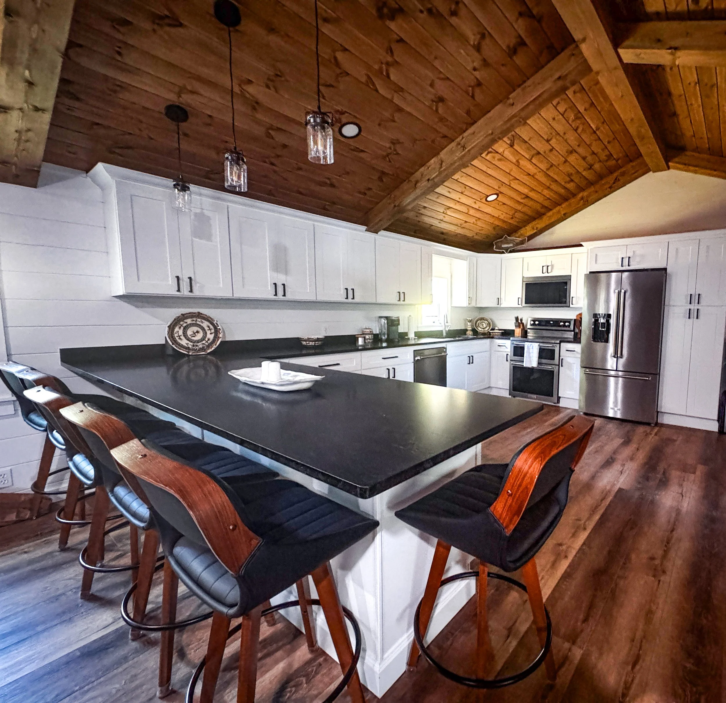 Modern kitchen with white cabinets, black countertops, stainless steel appliances, a large island with four bar stools, wood-paneled ceiling, and hardwood floors.