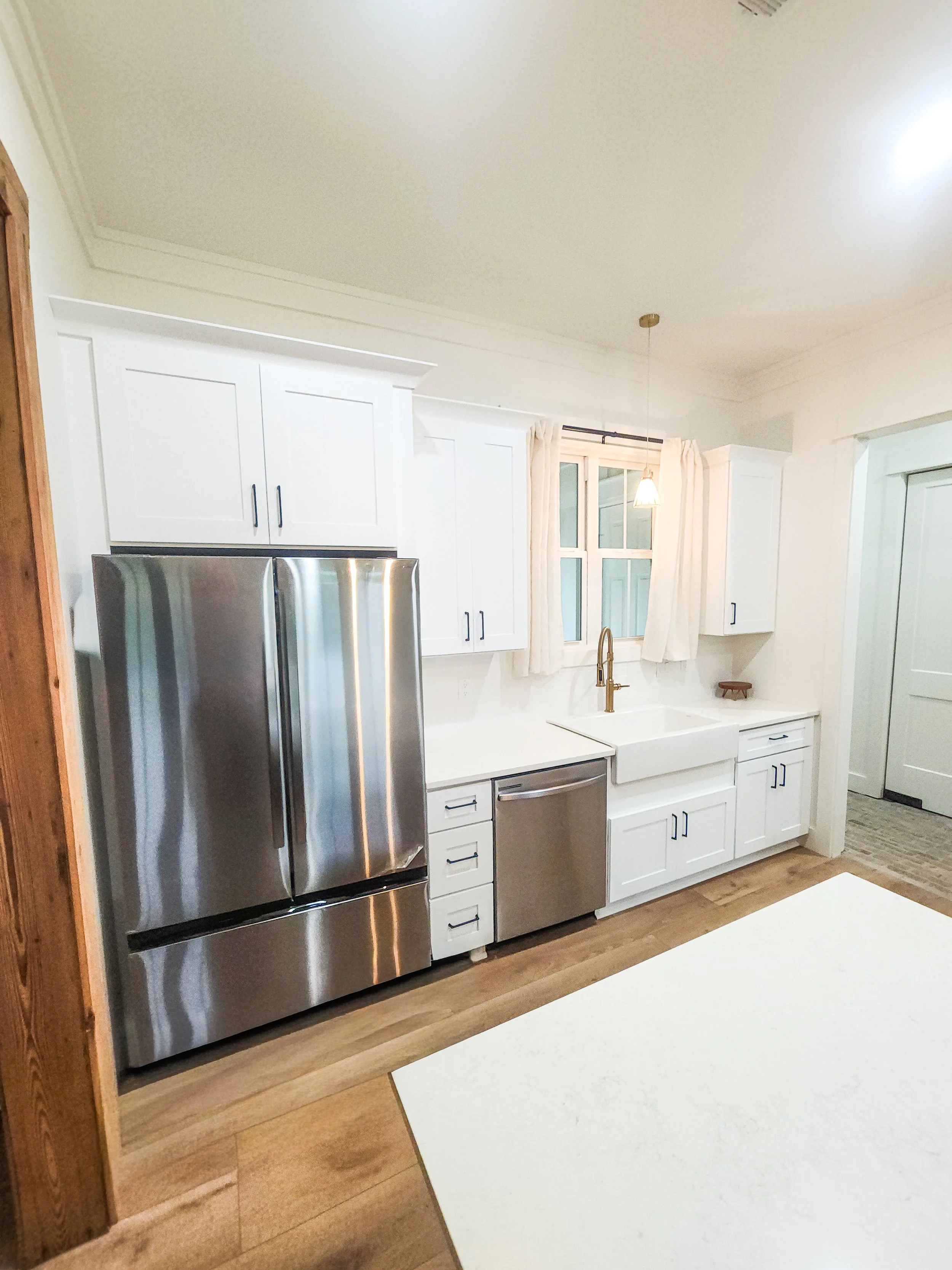 Modern kitchen with white cabinetry, stainless steel refrigerator and dishwasher, farmhouse sink, wooden floors, small window with cream curtains, and pendant light.