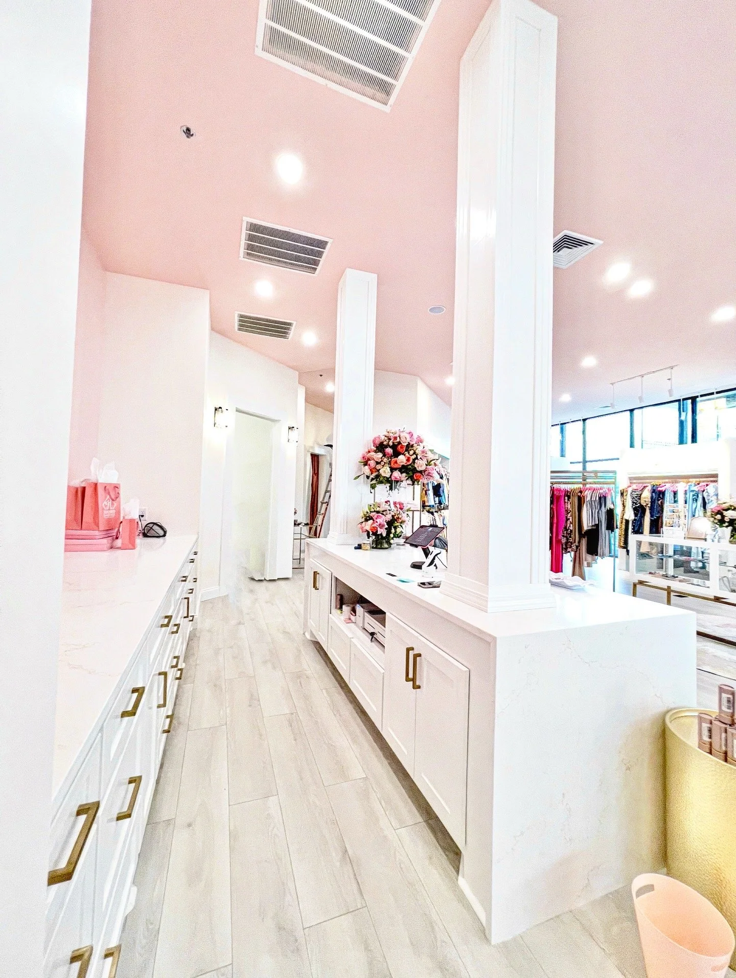 Interior of a retail store with pink ceiling, white cabinetry, and a floral display on counter, clothing racks visible in background.
