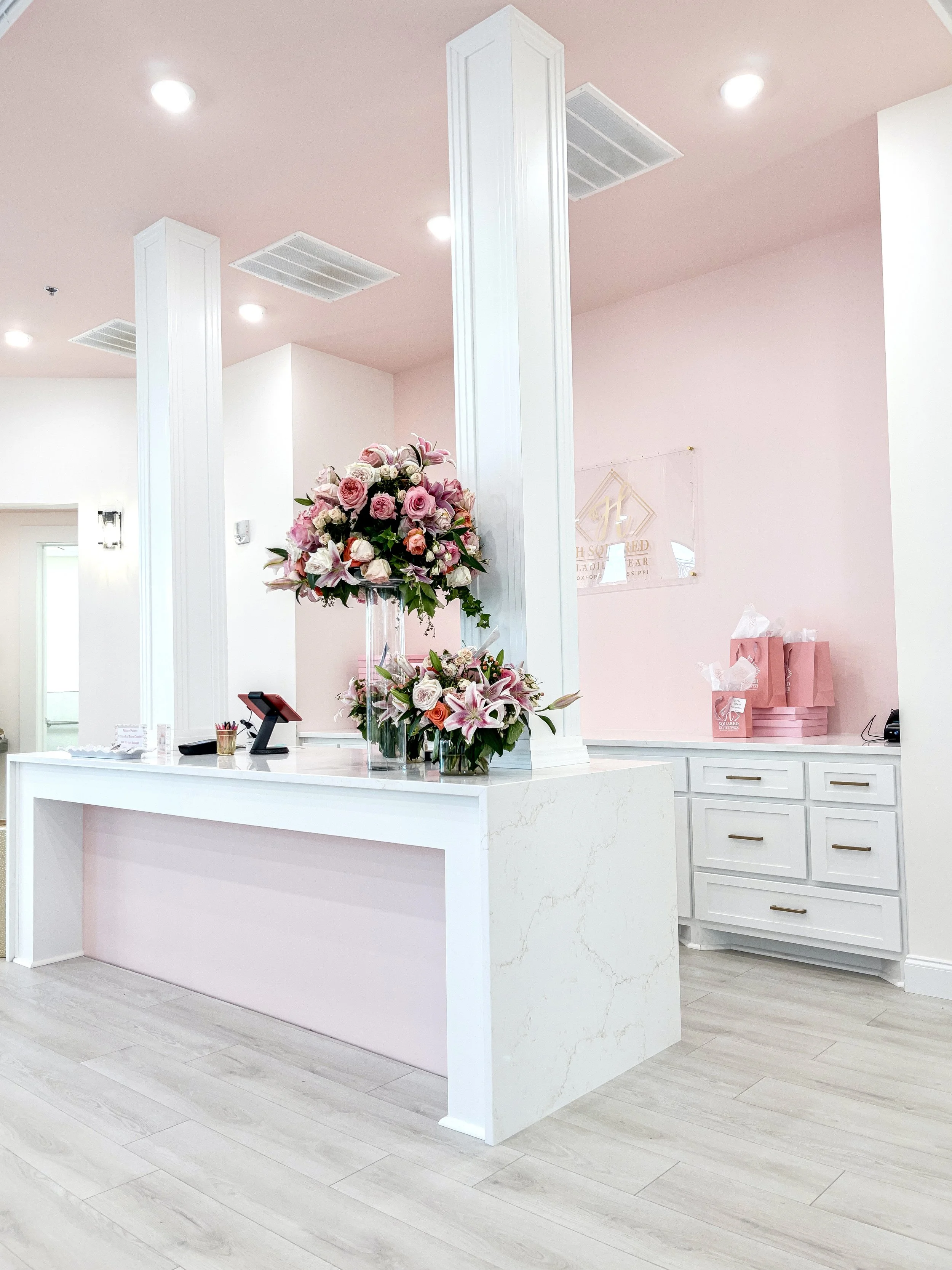 Interior of a reception desk with a large bouquet of pink and white flowers, pink gift bags on a counter, and a light pink wall in the background.