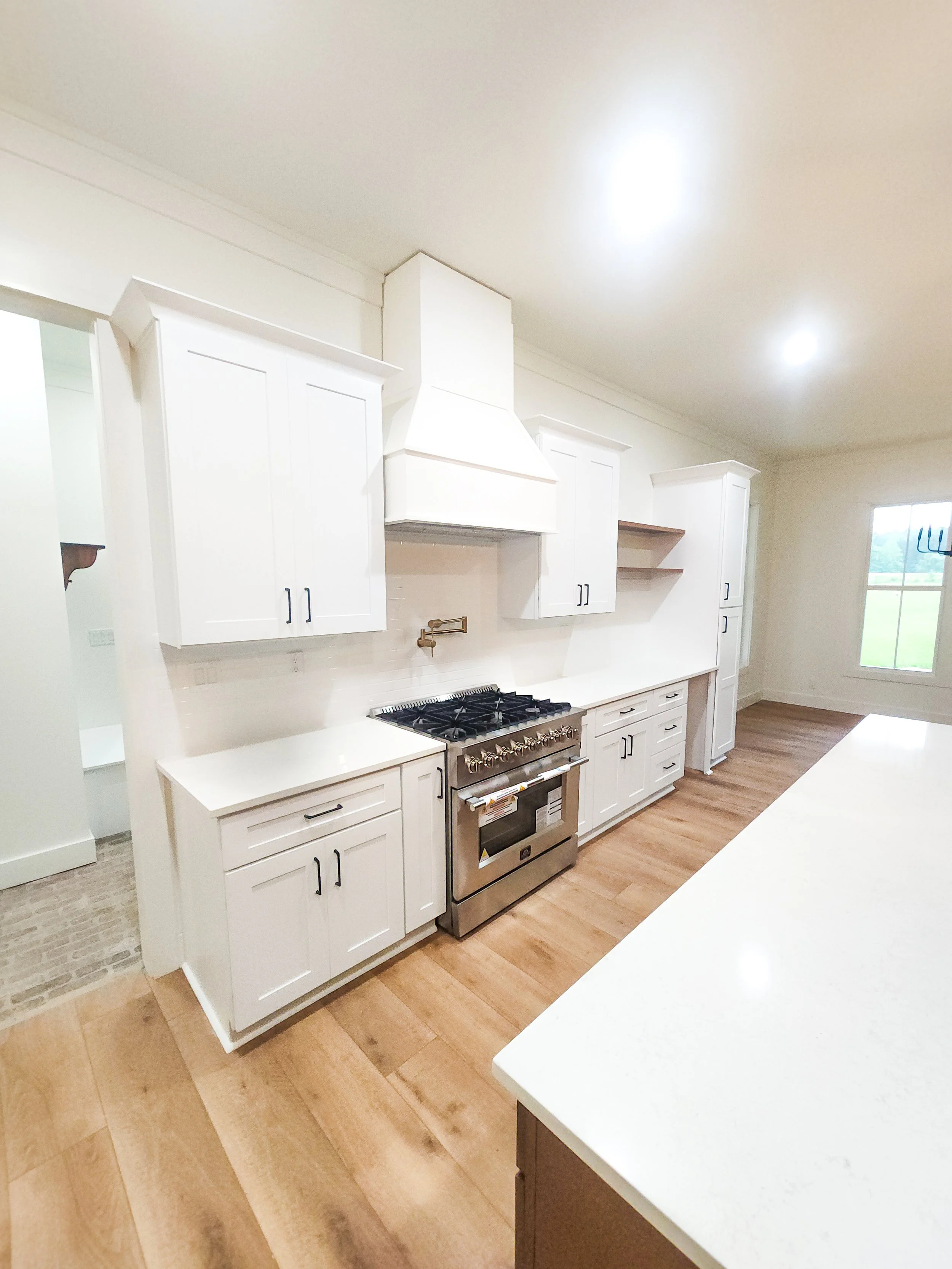 White kitchen with wooden flooring, white cabinets, a stainless steel oven and stove, pendant lights, and a window showing outdoor scenery.