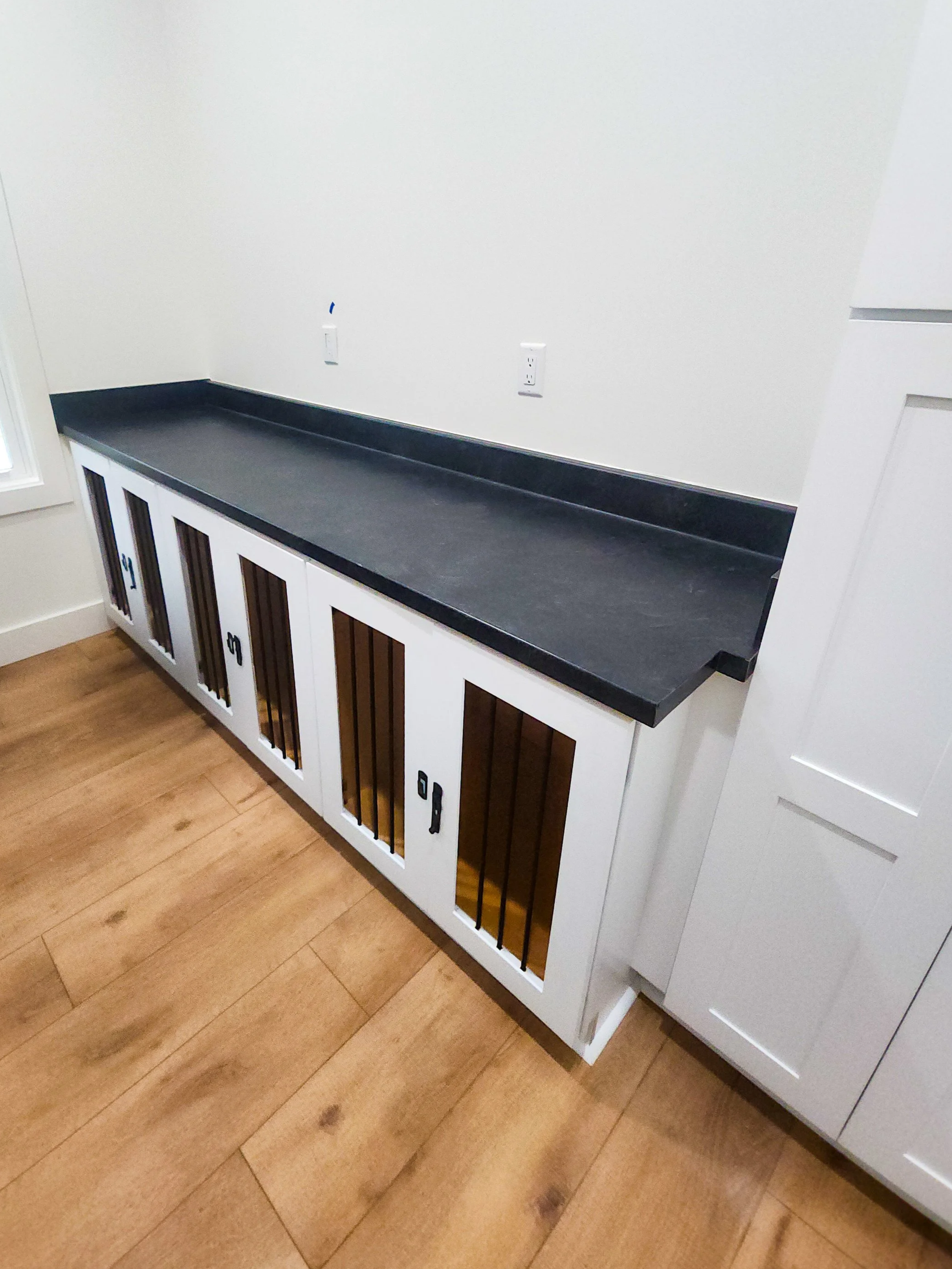 A white cabinet with glass door panels and black bars on a wooden floor, set against a white wall with electrical outlets.