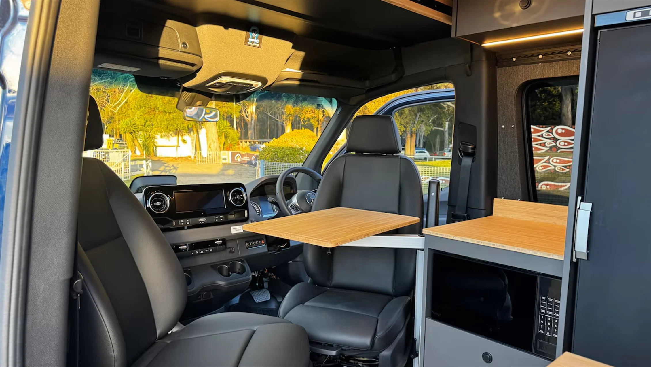 Interior of a converted van with black leather seats, a wooden fold-out table, and a kitchenette area with a microwave, all illuminated by natural light.